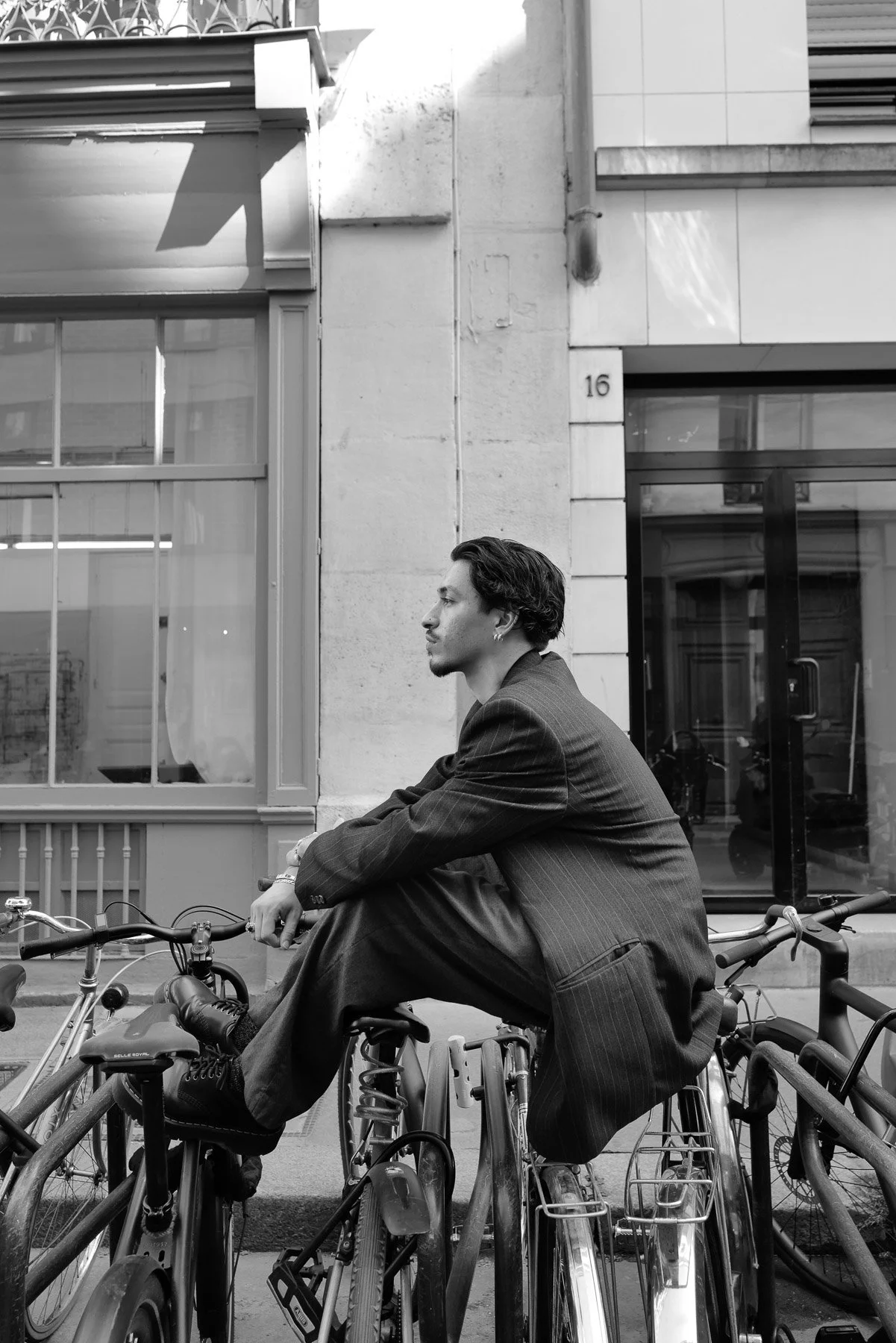 A man in a pinstripe suit sitting on a bicycle in front of a building with a large window and a door, looking to the side, in black and white.