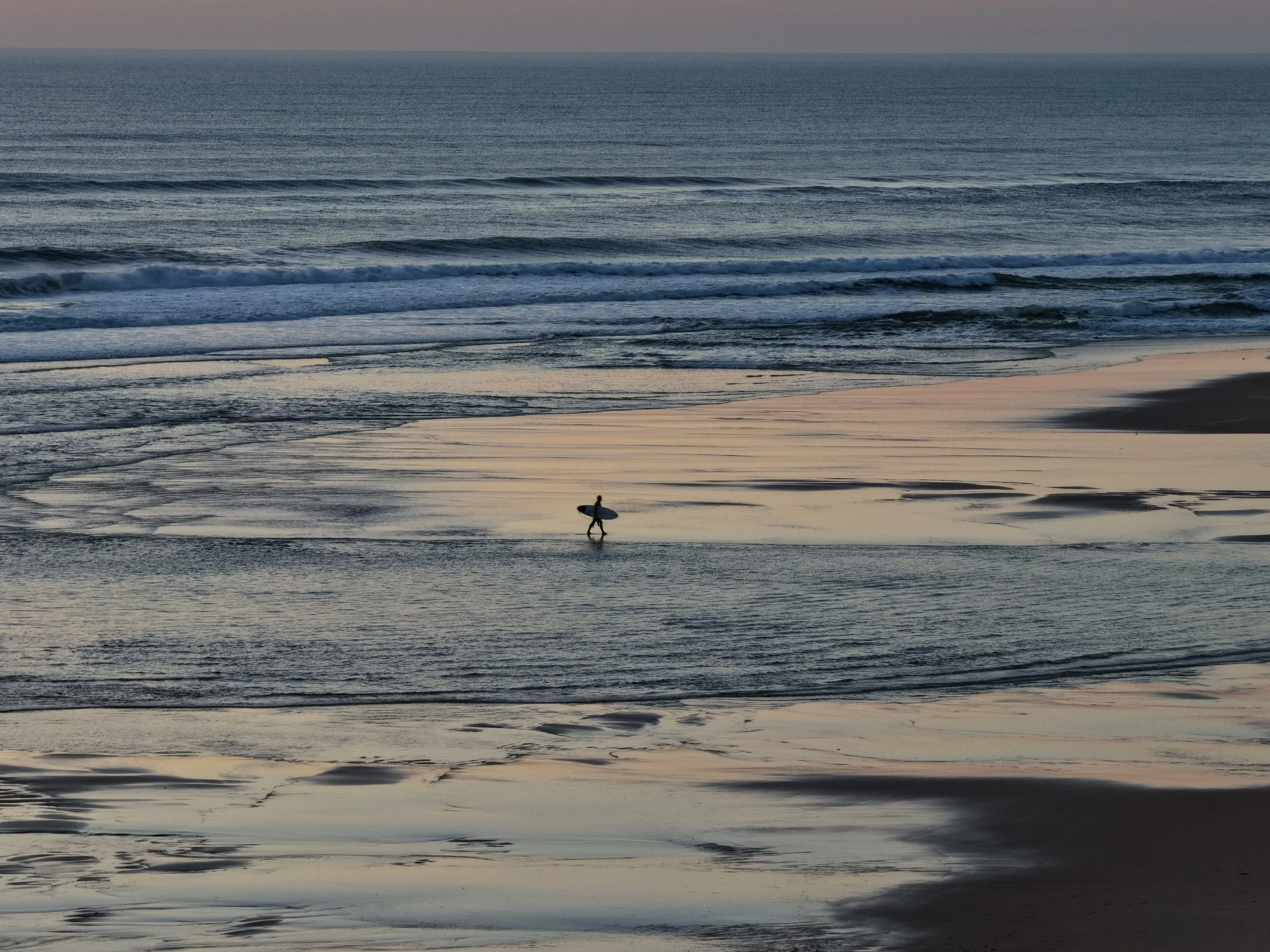 A person carrying a surfboard walking on a wet beach with gentle waves during sunset or dawn.
