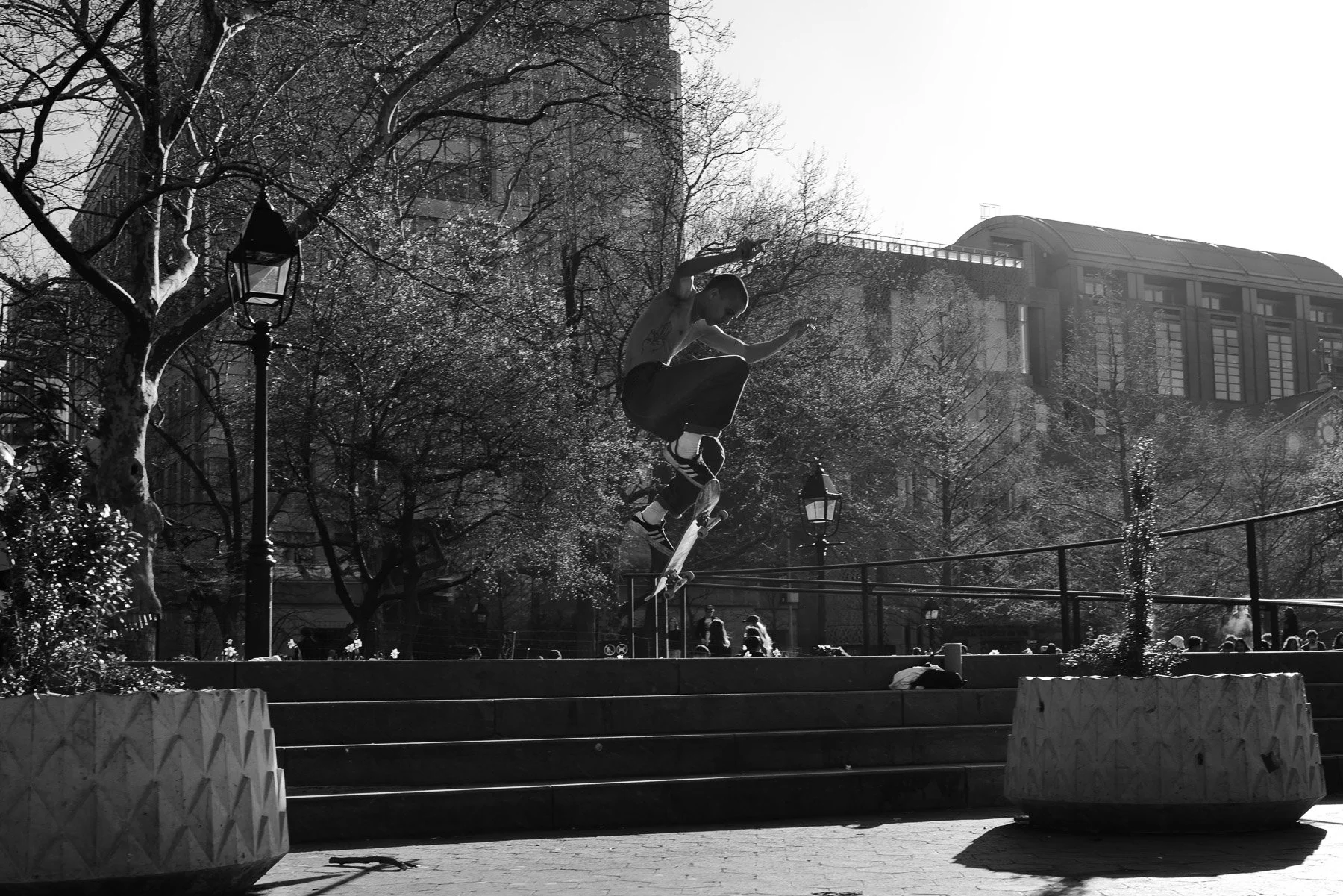 A young person performing a skateboarding trick in an urban park, captured in black and white with city buildings and leafless trees in the background.