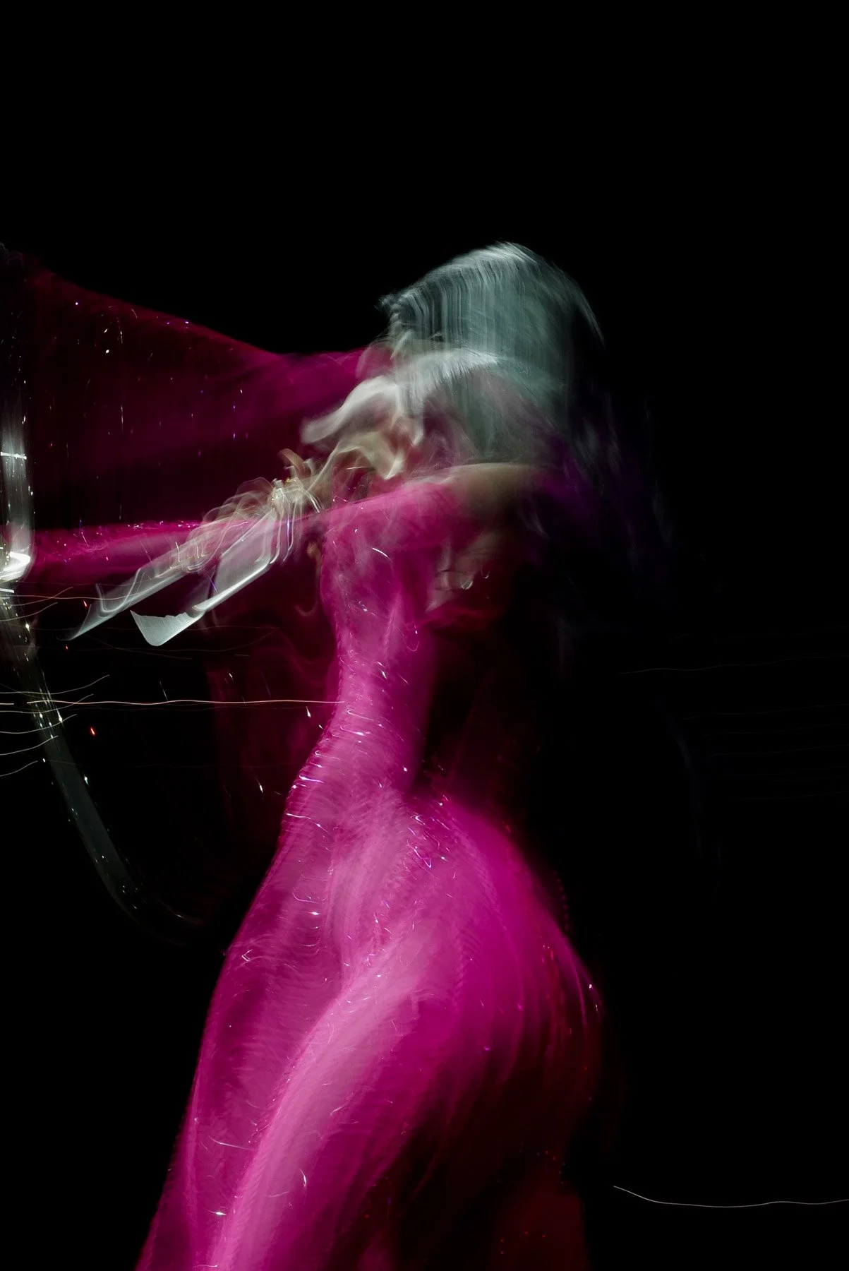 A person wearing a vibrant pink dress with long sleeves is blowing smoke or vapor through a glass pipe, with the smoke swirling around her head. She has short hair and is set against a dark background.