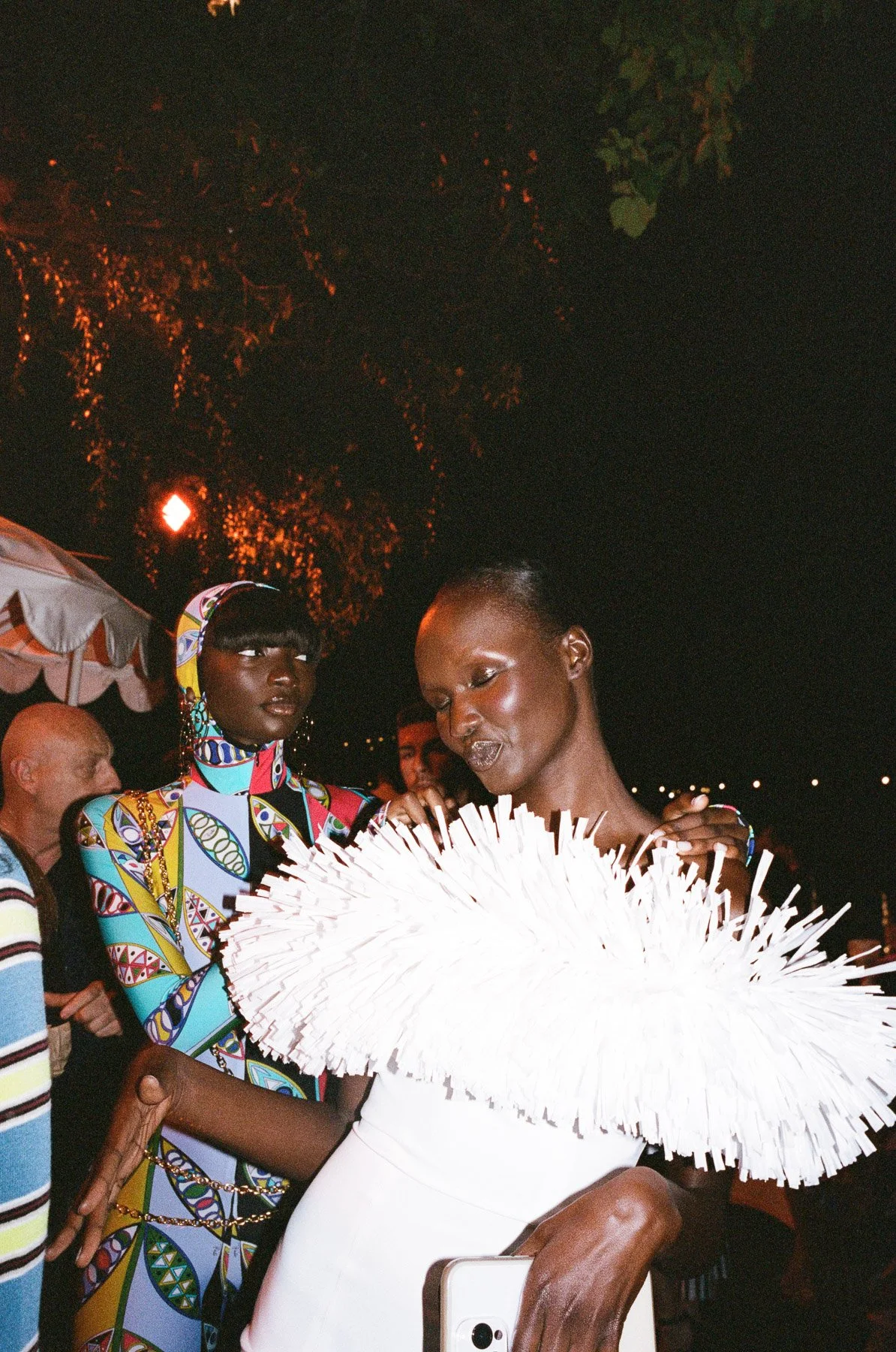 Two women at night, one wearing colorful patterned clothing and the other in white holding a large white feathered fan, with people and trees in the background.