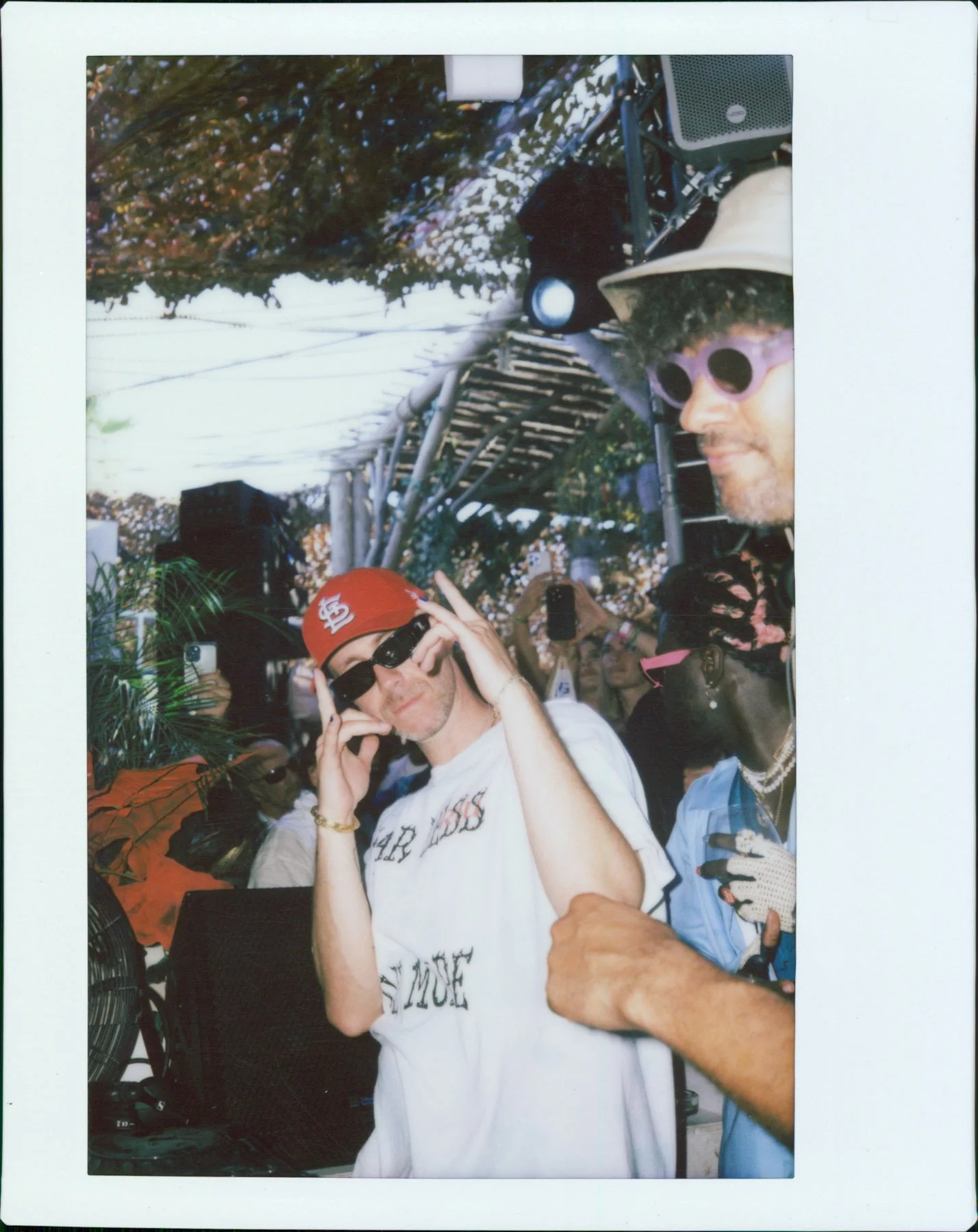 Two people taking a selfie together amidst a crowd at an outdoor event. One person wears a red baseball cap, sunglasses, and a white t-shirt. The other person wears purple sunglasses, a hat, and a blue shirt with jewelry. The background shows a ladde