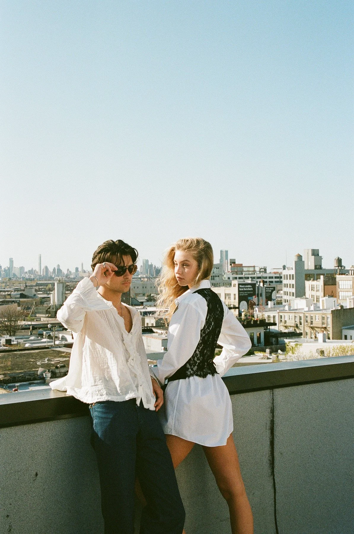 Two young women stand on a rooftop with a city skyline behind them. One woman wears sunglasses, a white blouse, and blue jeans. The other woman has long blonde hair, a white dress, and a black vest.