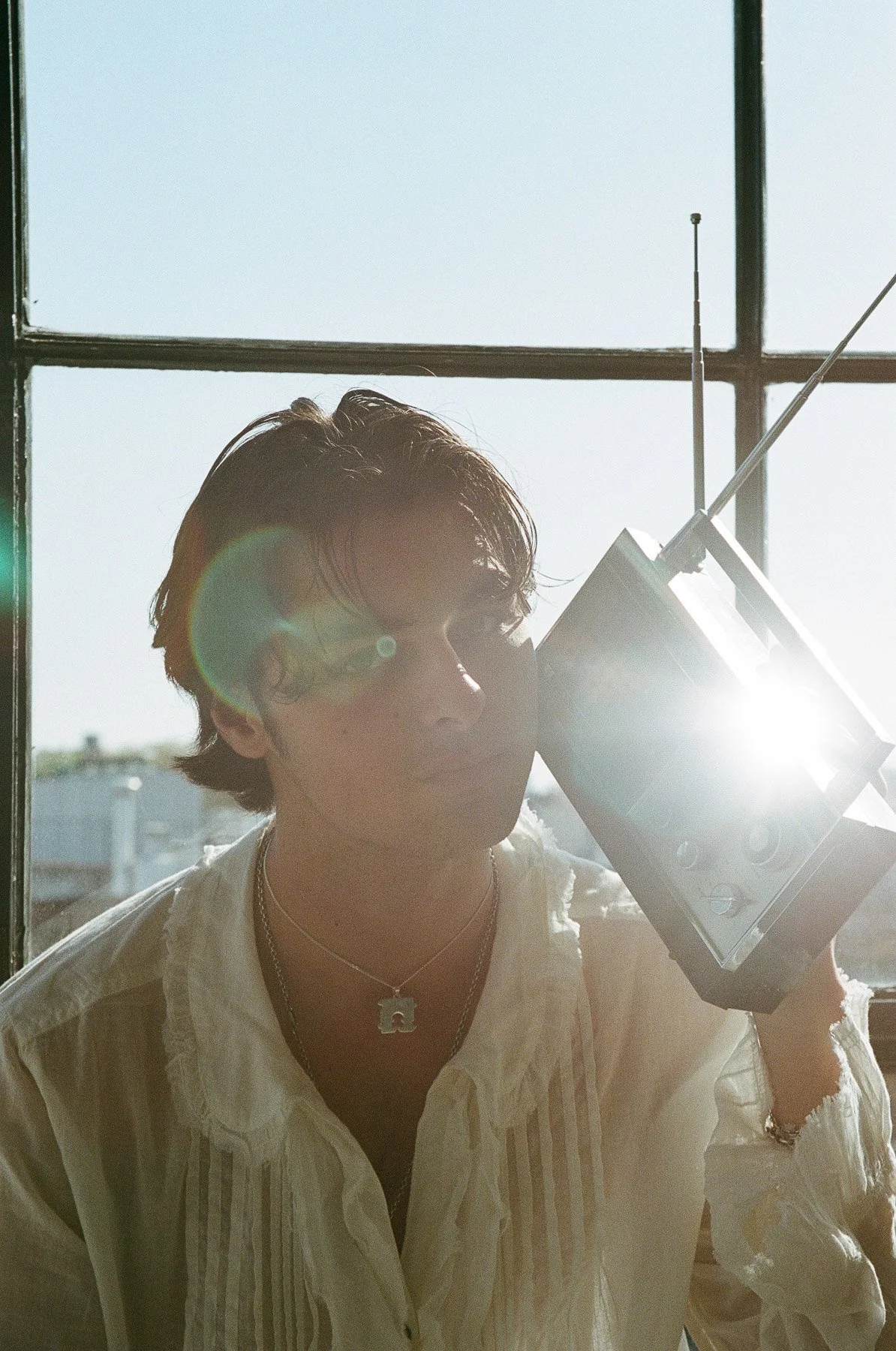 A person with wavy hair is holding a vintage radio to their ear, standing near a window with sunlight streaming in, creating a lens flare.