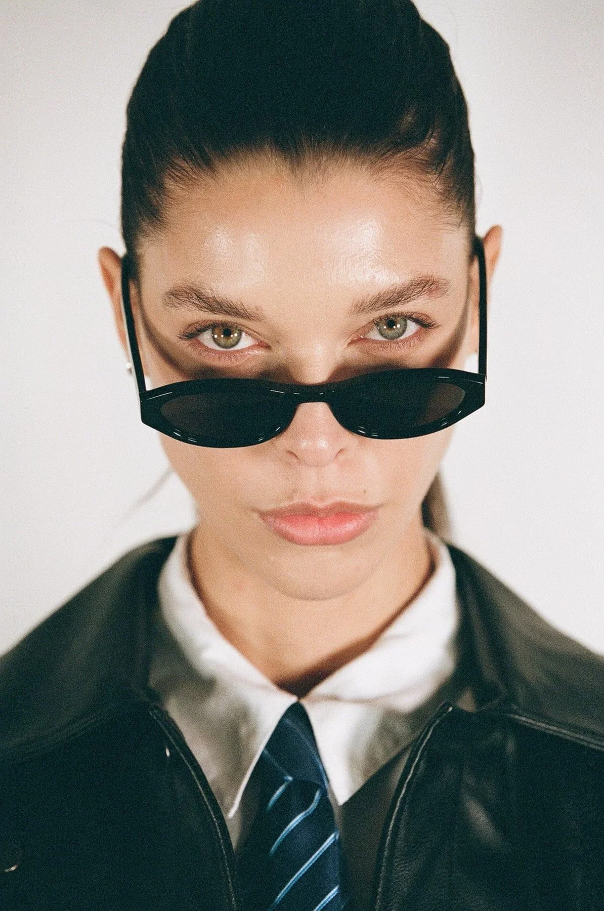 Close-up of a woman with dark hair pulled back, wearing black sunglasses, a white collared shirt, a blue striped tie, and a black leather jacket, against a white background.