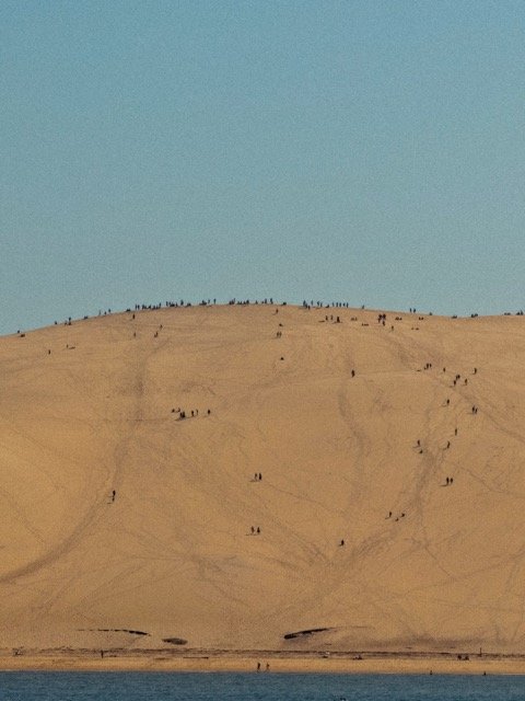 A large sandy hill with many small figures walking and gathering on the slope, and a few at the base near a body of water, under a clear blue sky.