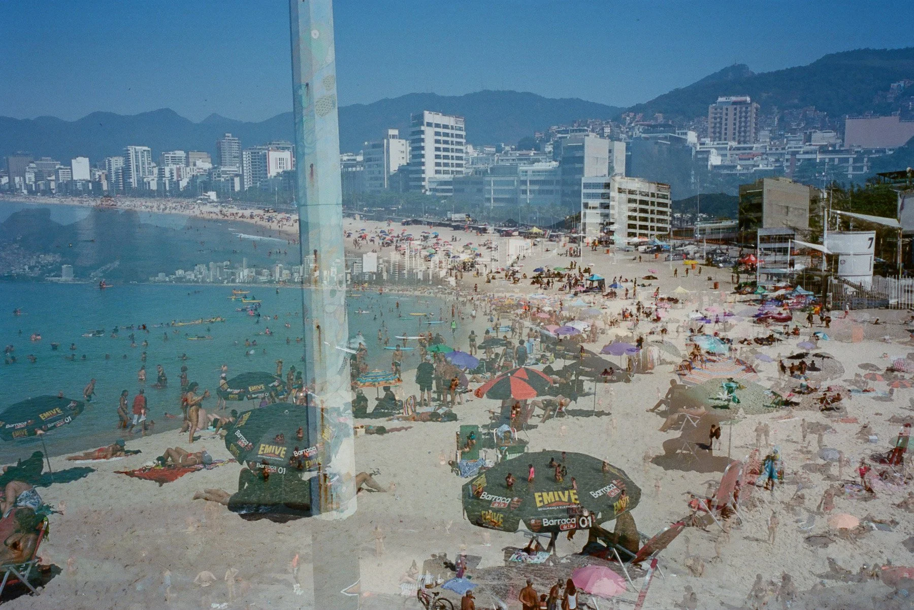 A crowded beach with many people, umbrellas, and chairs, with tall buildings and mountains in the background, viewed through a glass window.