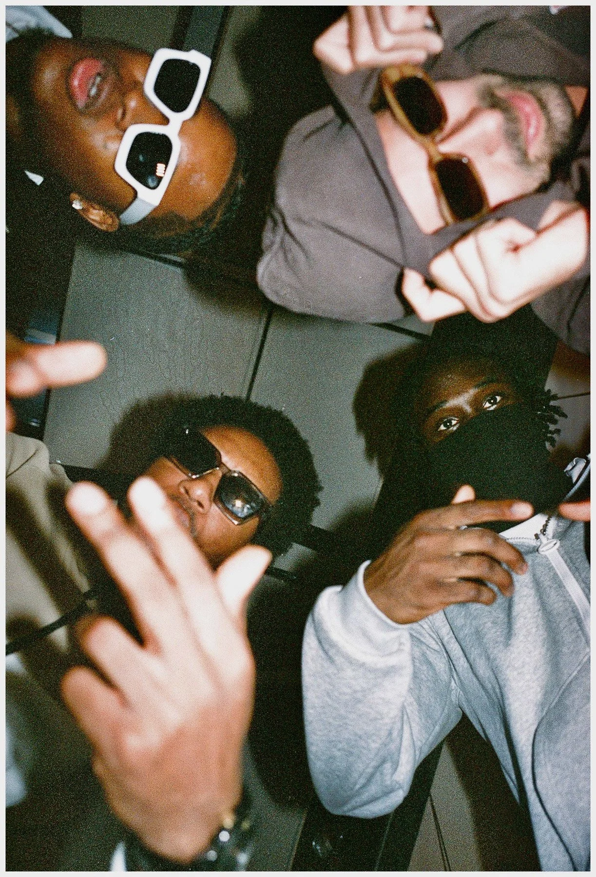 Four friends lying on a black tiled floor, wearing sunglasses and casual clothing, making hand gestures and looking up at the camera.
