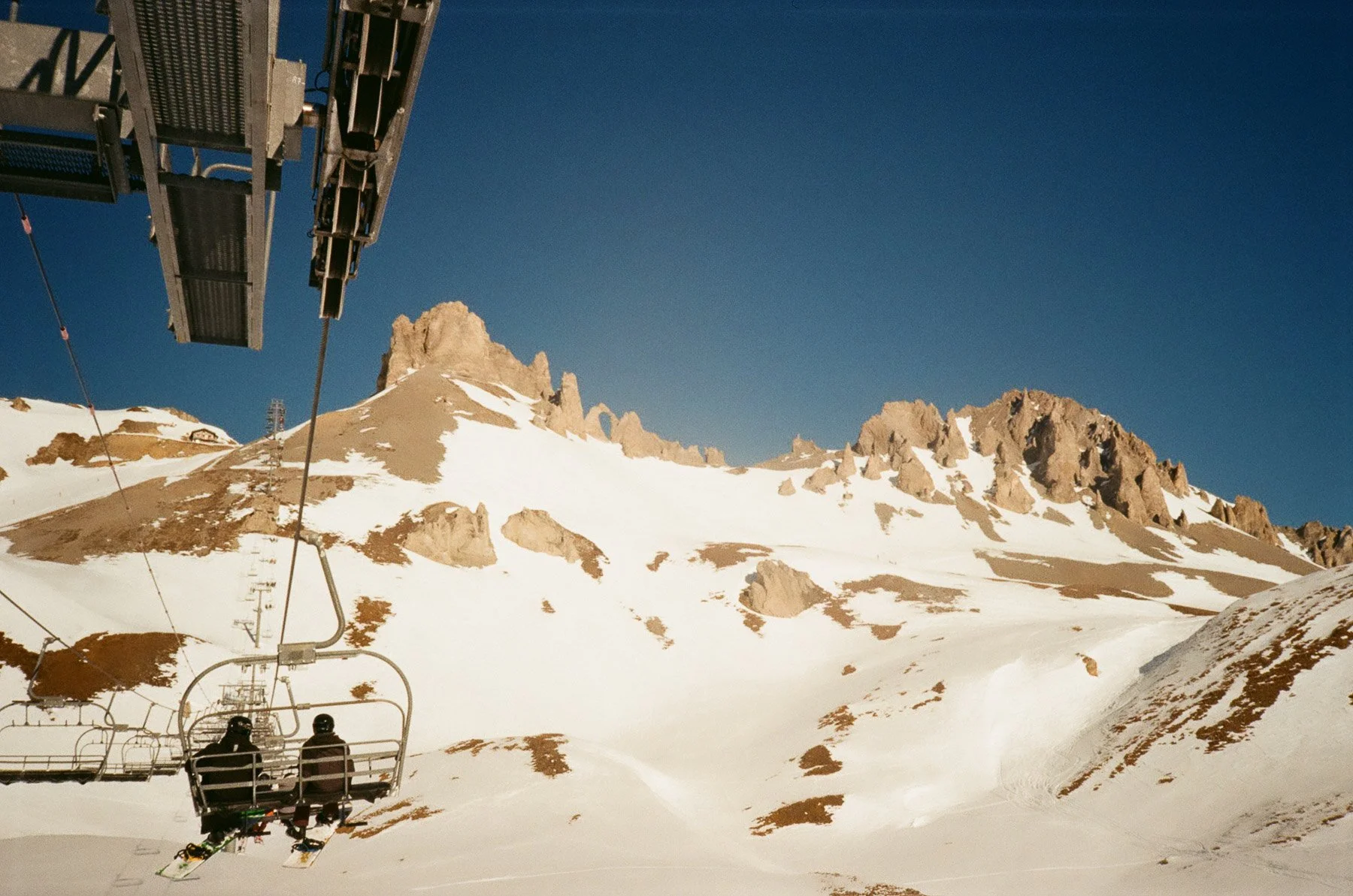Ski lift going up a snowy mountain with rocky peaks in the background under a clear blue sky.
