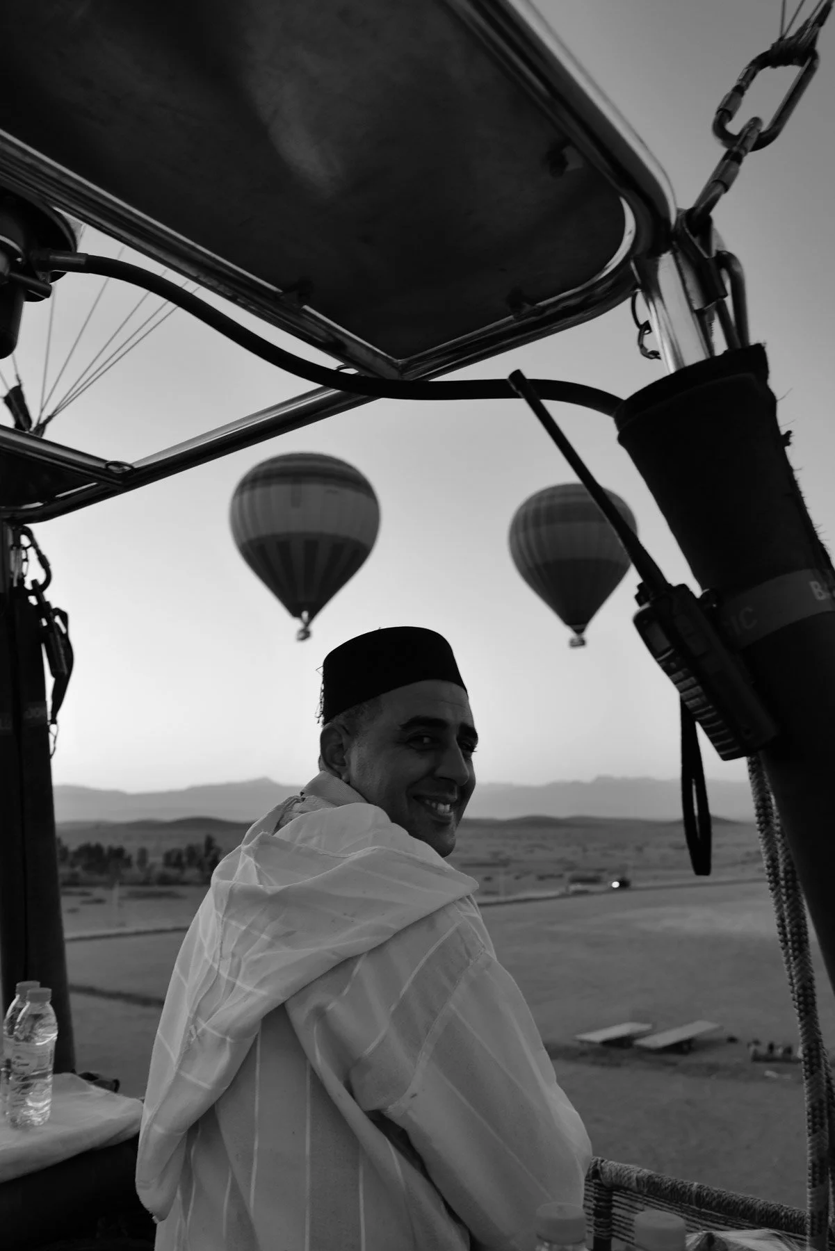 A man smiling and wearing a hat, sitting in a hot air balloon basket with hot air balloons in the background.