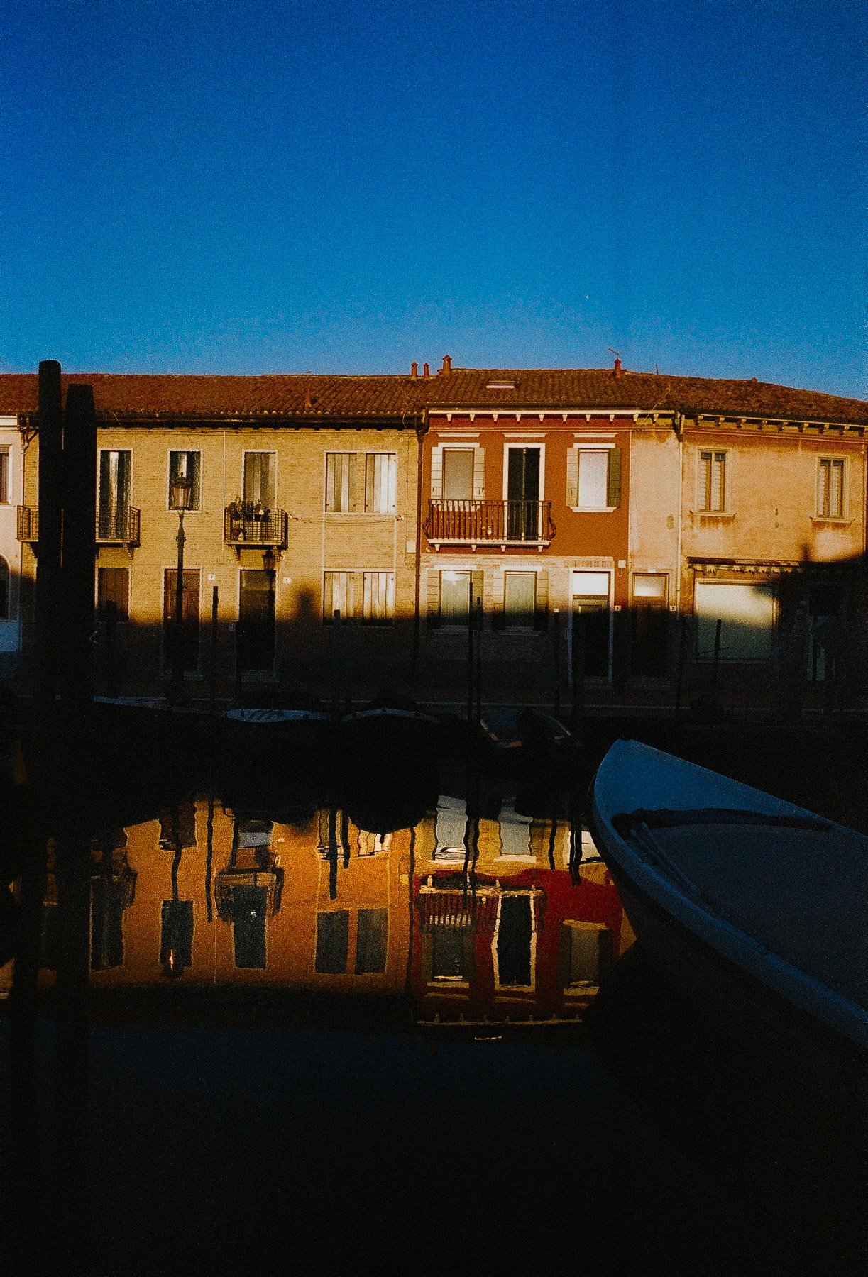 Reflected image of a row of buildings with balconies, under a clear blue sky, with boats in the foreground on the water.