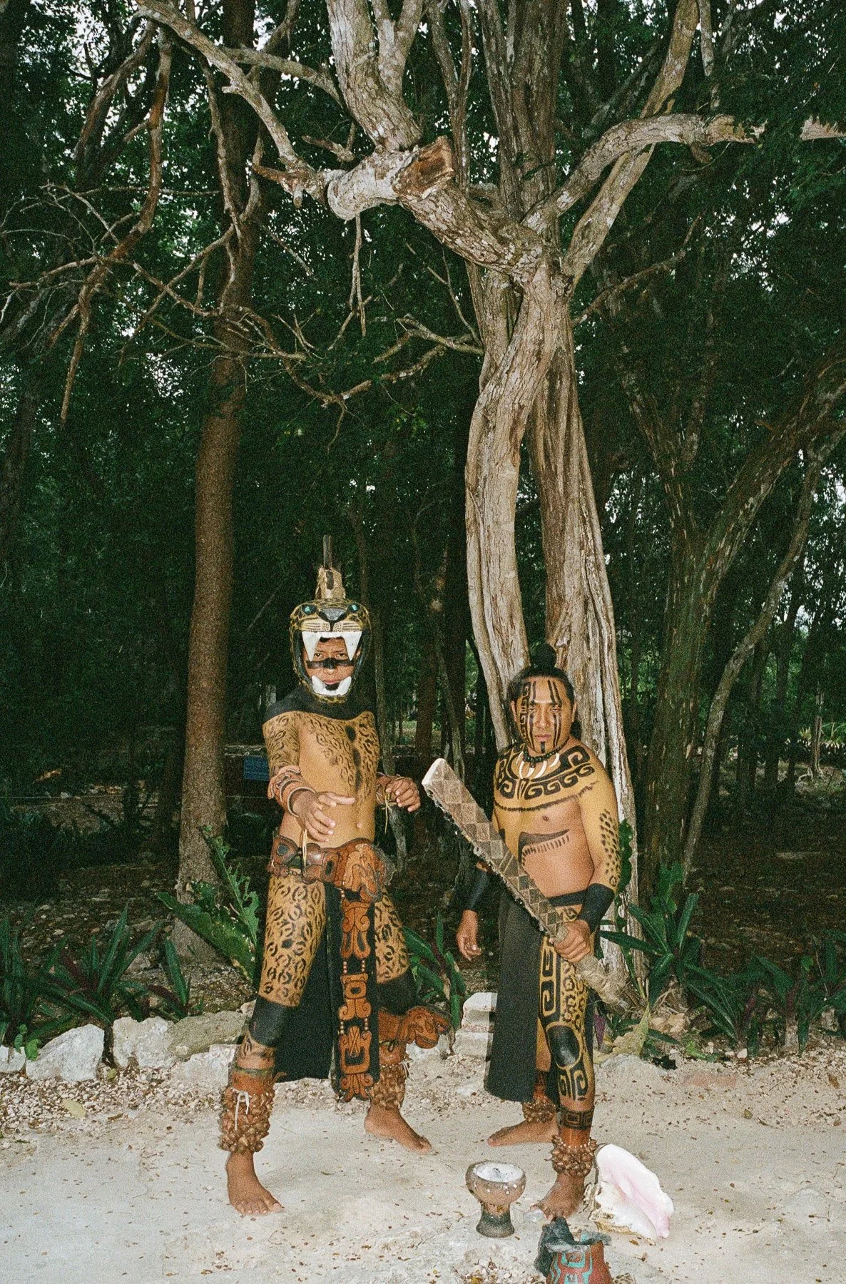 Two men dressed in traditional tribal costumes with face paint and accessories standing in a forested area near a large tree, holding a weapon and a staff, with ritual objects placed on the ground in front of them.