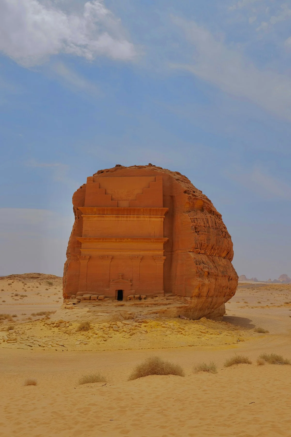 Large ancient sandstone building with carved steps and details on a desert landscape under a partly cloudy sky.