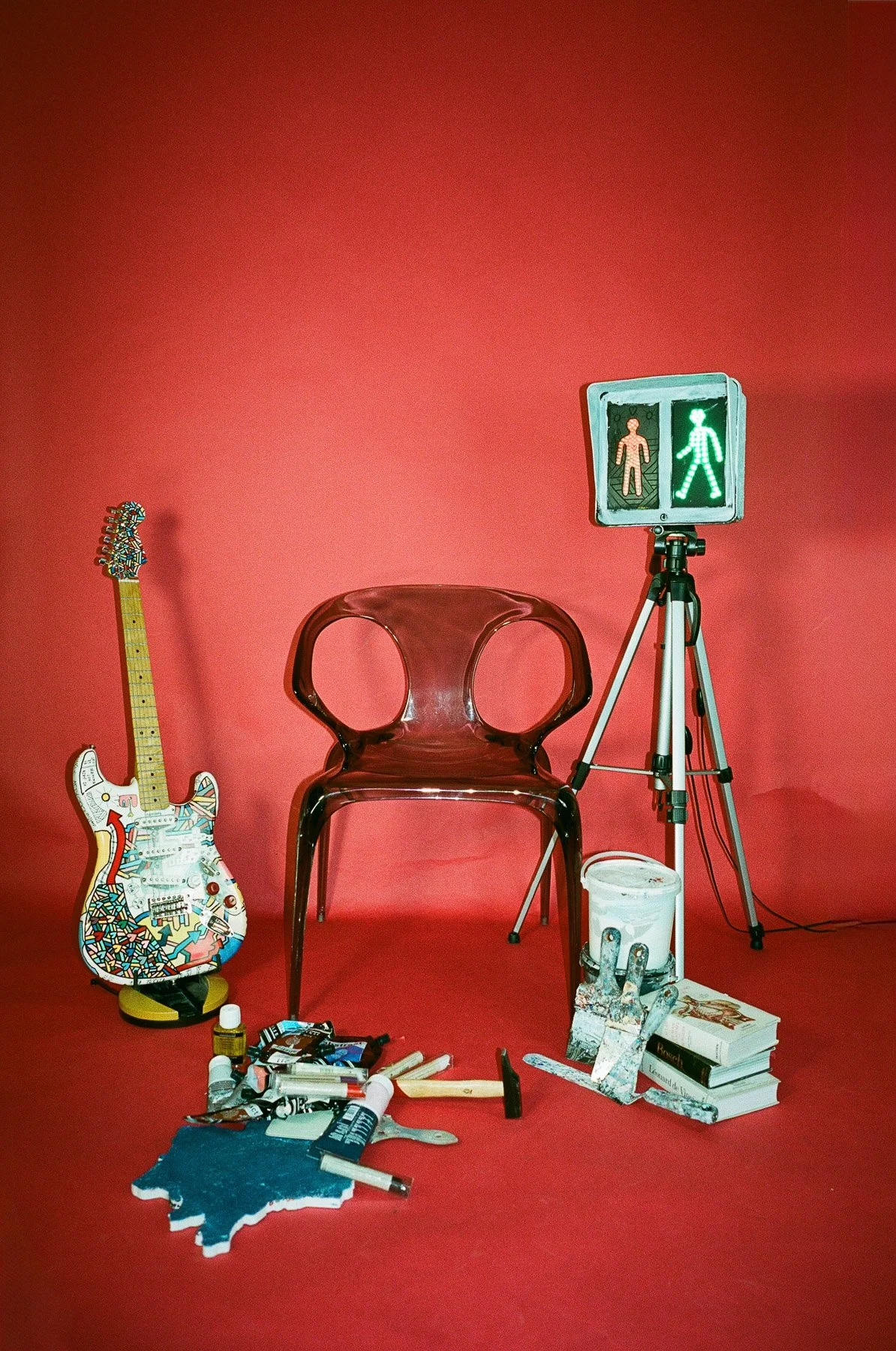 A red background with a guitar decorated with colorful patterns on a stand, a transparent plastic chair, a tripod with a green pedestrian crossing signal, art supplies, books, and a white bucket in a studio or art space.