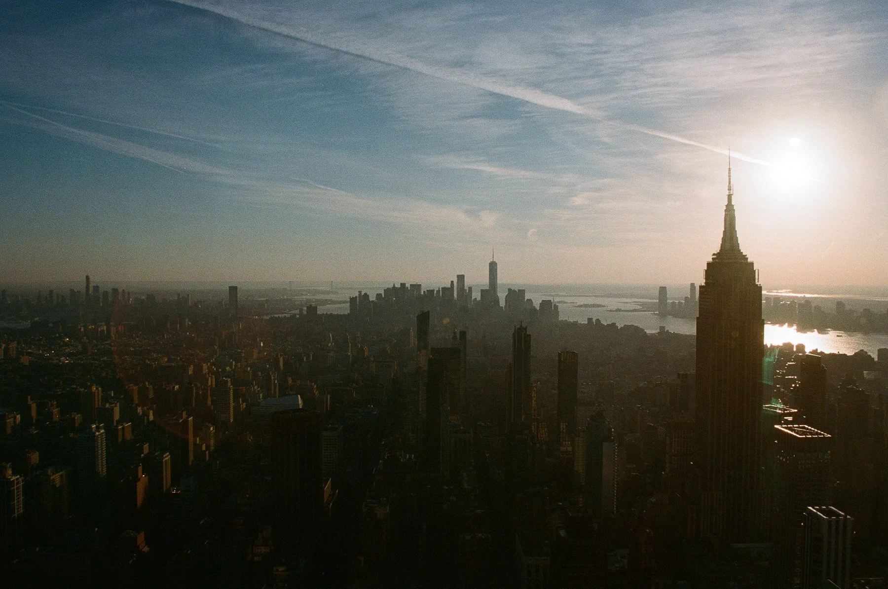 A panoramic view of New York City skyline at sunset, featuring the Empire State Building prominently in the foreground and the cityscape extending towards the horizon with water bodies and other skyscrapers visible in the distance.