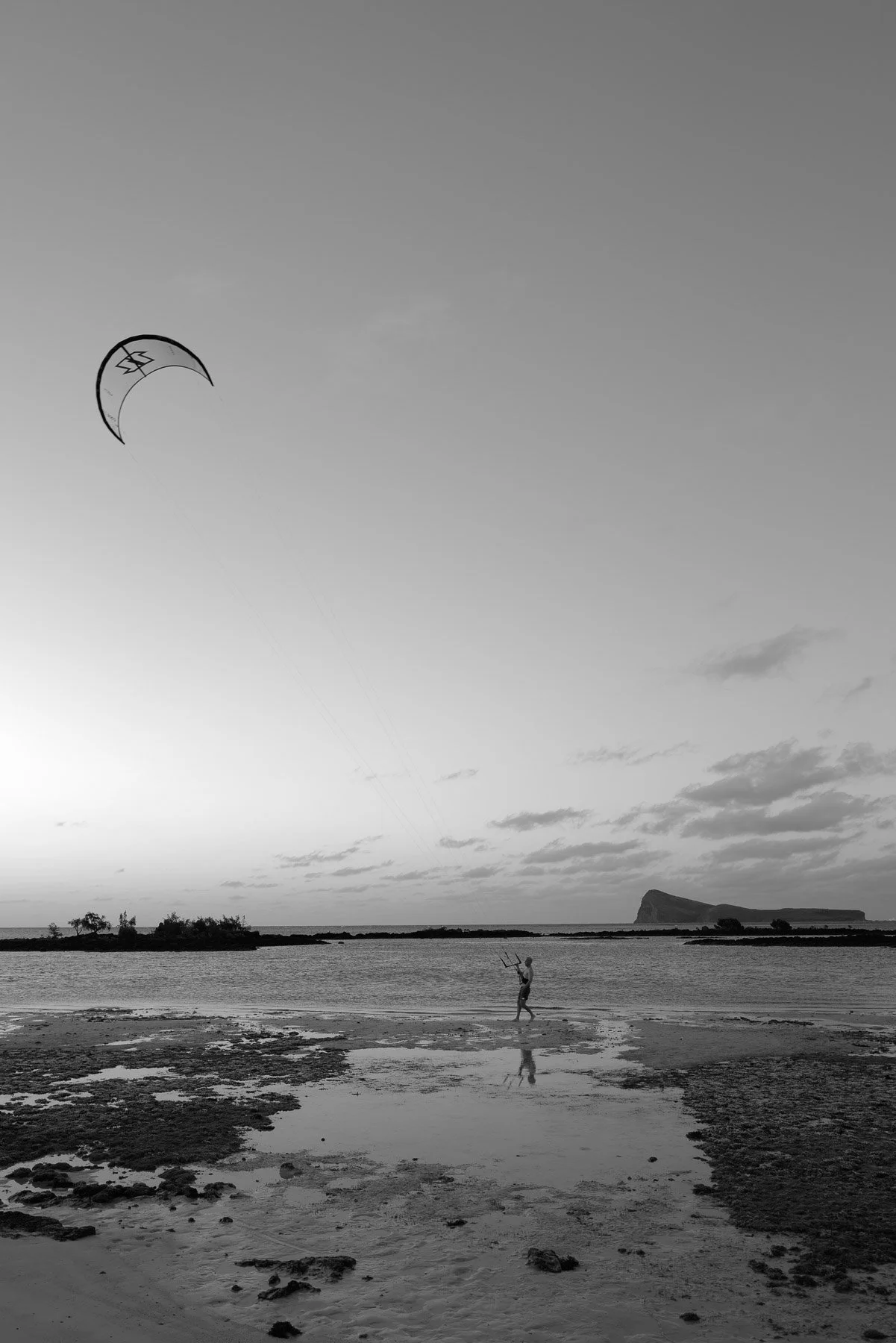 A black and white photo of a person on a beach flying a kite with the ocean and distant landmass in the background.