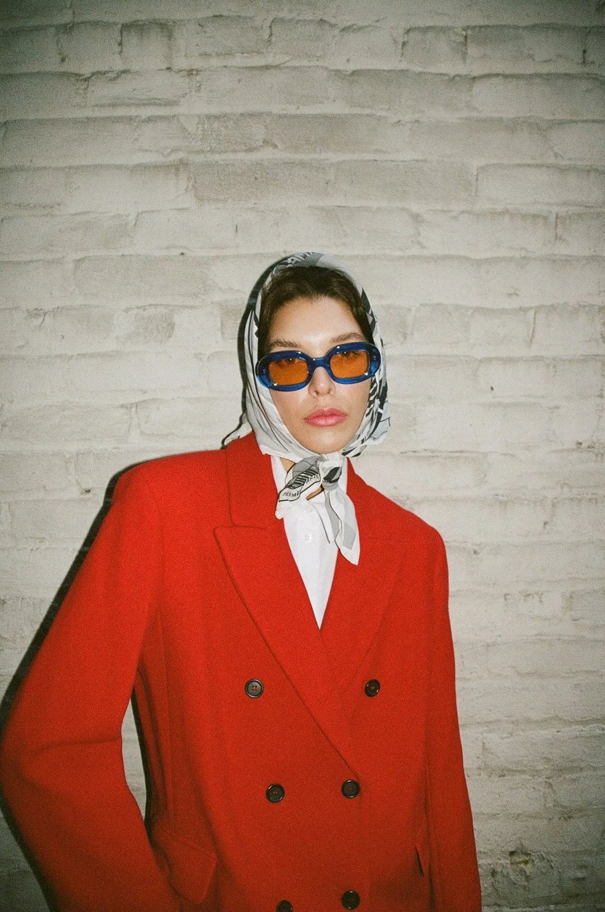 A woman wearing a red blazer, white shirt, patterned scarf, and blue sunglasses with orange lenses poses against a white brick wall.