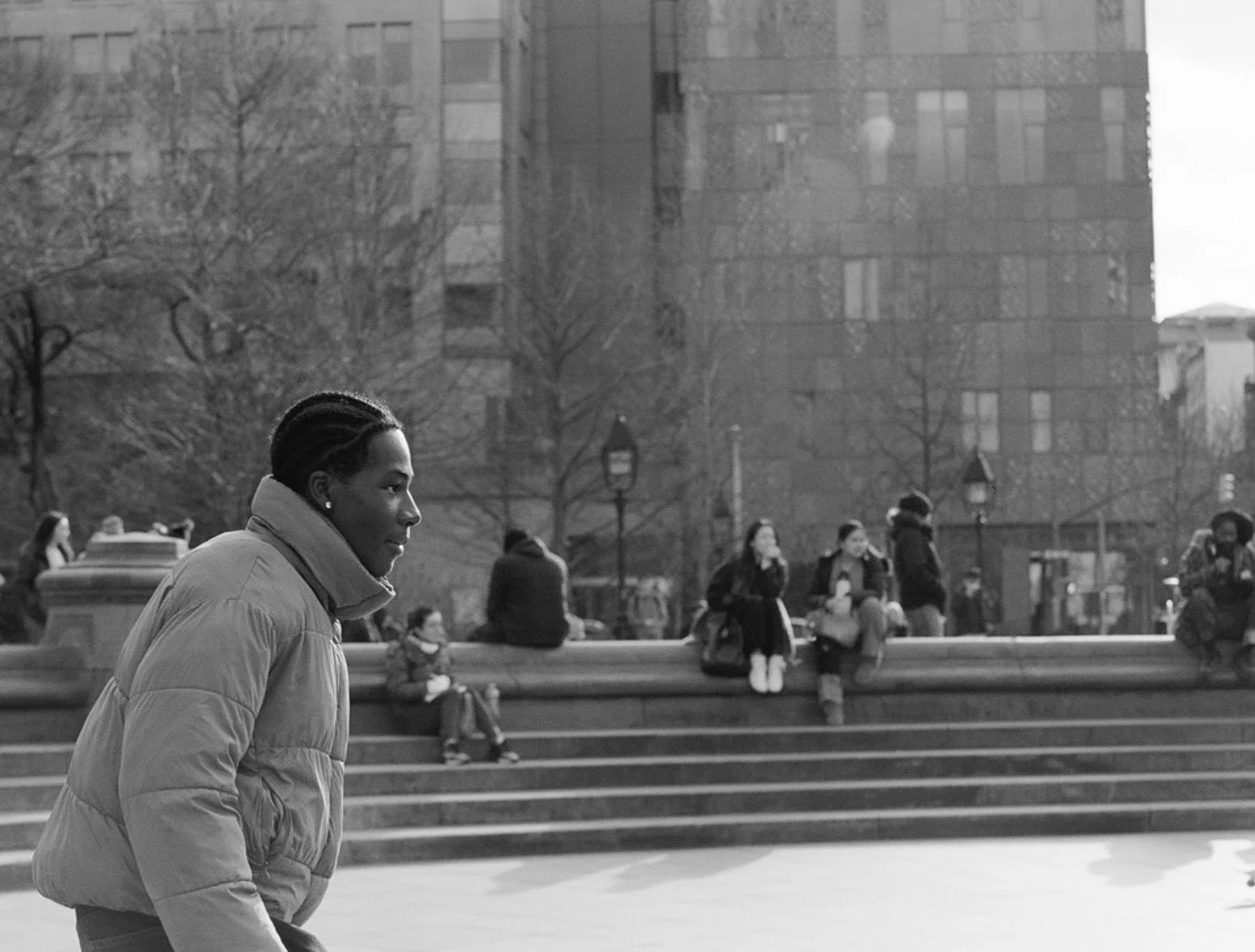 A woman with braided hair wearing a puffy jacket walking in an urban park with stairs, benches, trees, and people, black and white photograph.