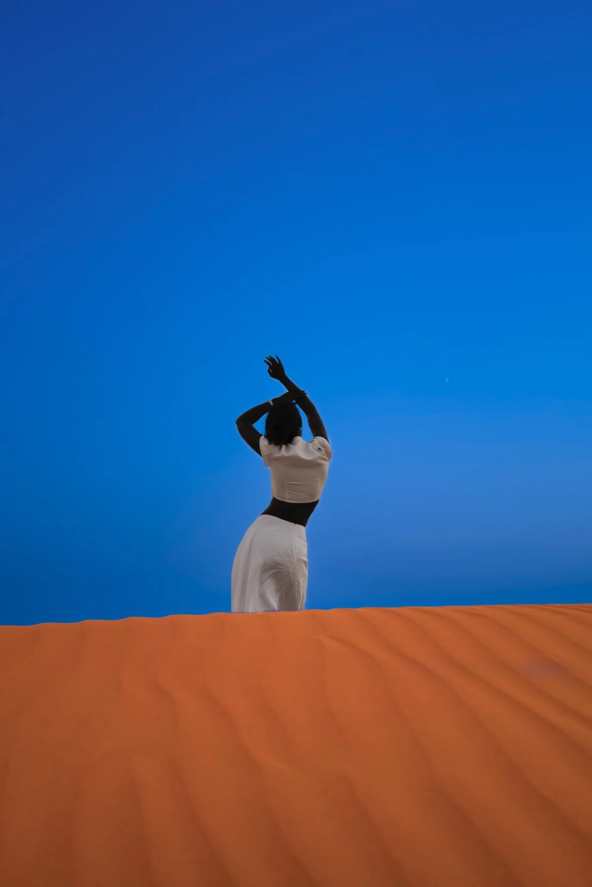 A silhouette of a woman standing on an orange surface against a clear blue sky, with her hands raised above her head.