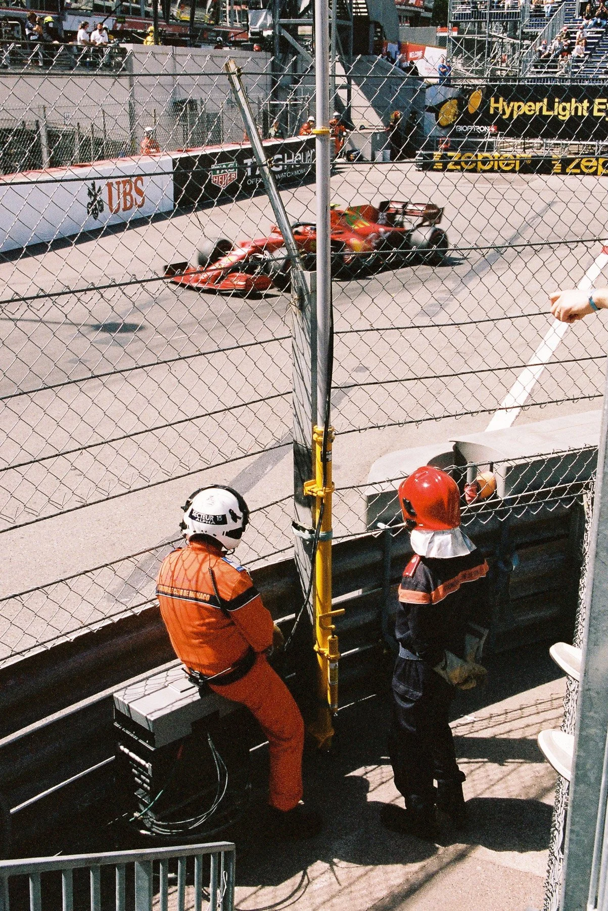A Formula 1 race car on a race track, with two officials and a security person watching from the side. The scene is set during a race event with spectators overhead and branding advertisements along the fence.