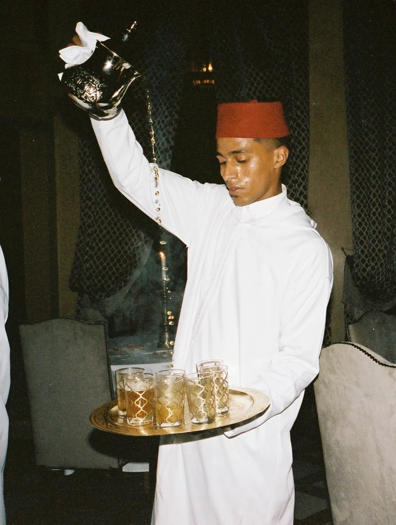 A waiter in traditional Moroccan attire holds a tray with several glasses of a golden-colored drink in a dimly lit restaurant.