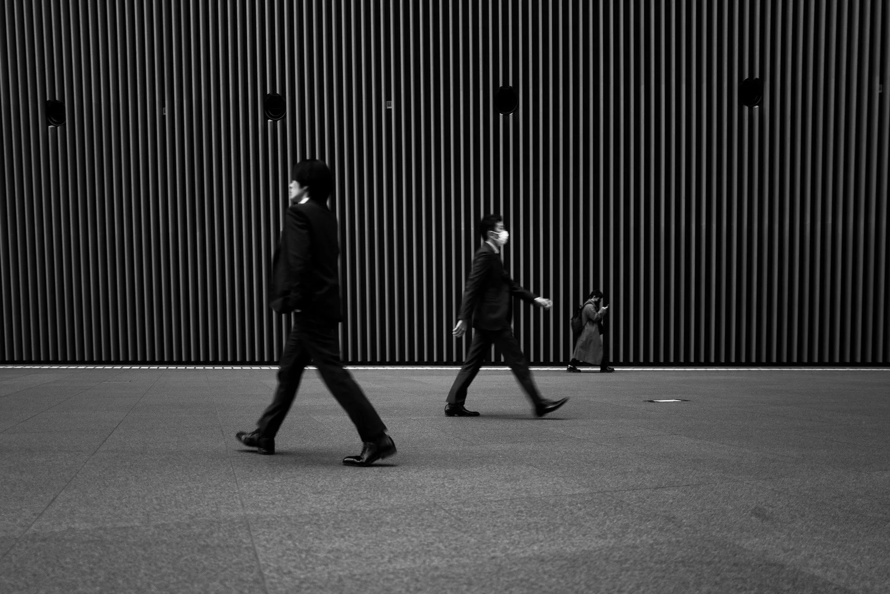 Black and white photo of three people walking in front of a textured wall. The person on the left is a man, the person in the middle is a woman with a face mask, and the person on the right is a woman also with a face mask. All are dressed in formal 