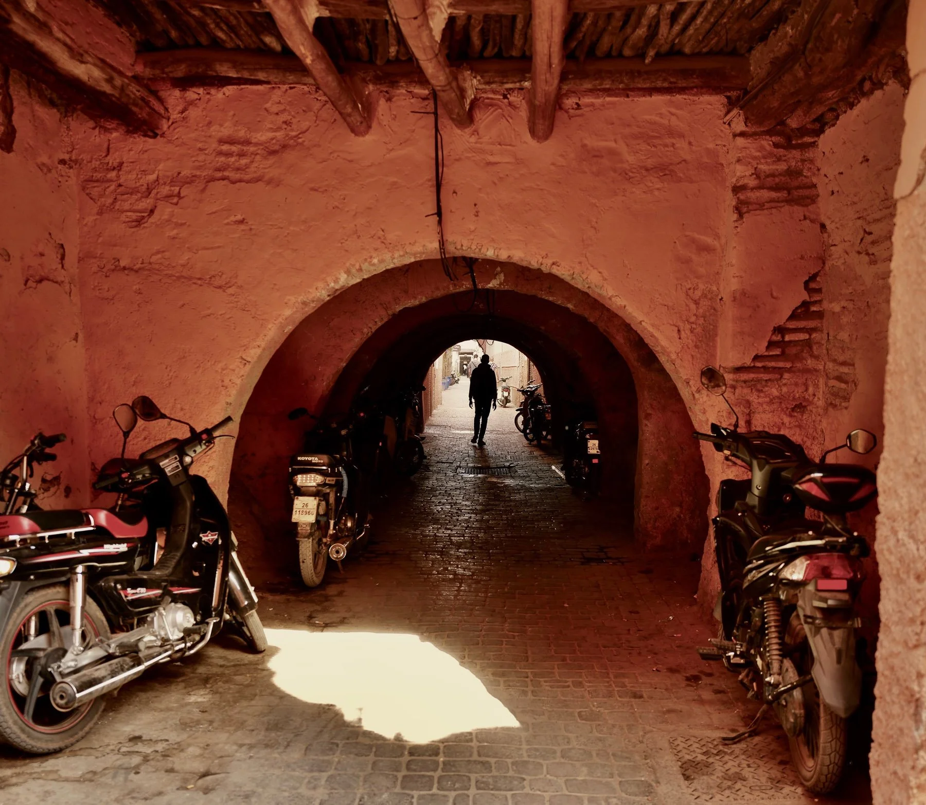 View through a brick archway in a Mediterranean or historical setting, showing parked motorcycles on both sides, a cobblestone street beyond, and a person walking in the distance.