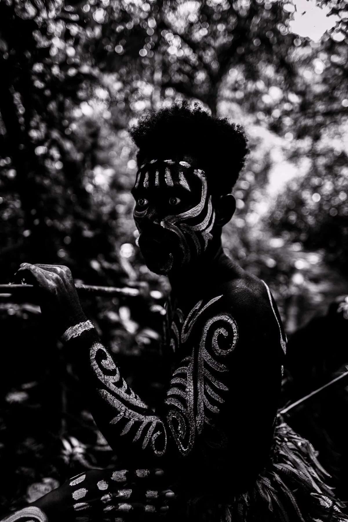 Person with traditional face and body paint, sitting in a forest, holding a bow.