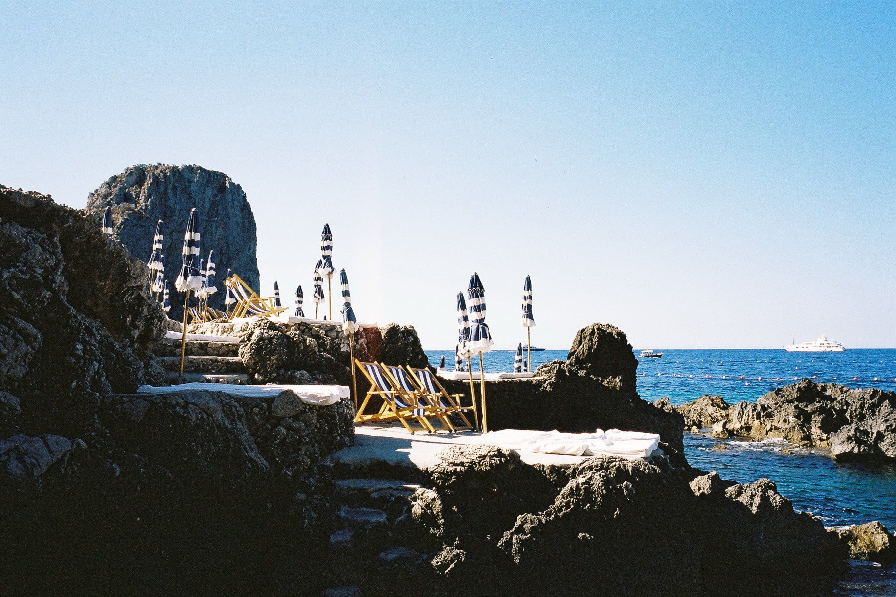 A rocky beach with blue and white striped umbrellas and lounge chairs, overlooking the ocean with boats and a large cliff in the background.