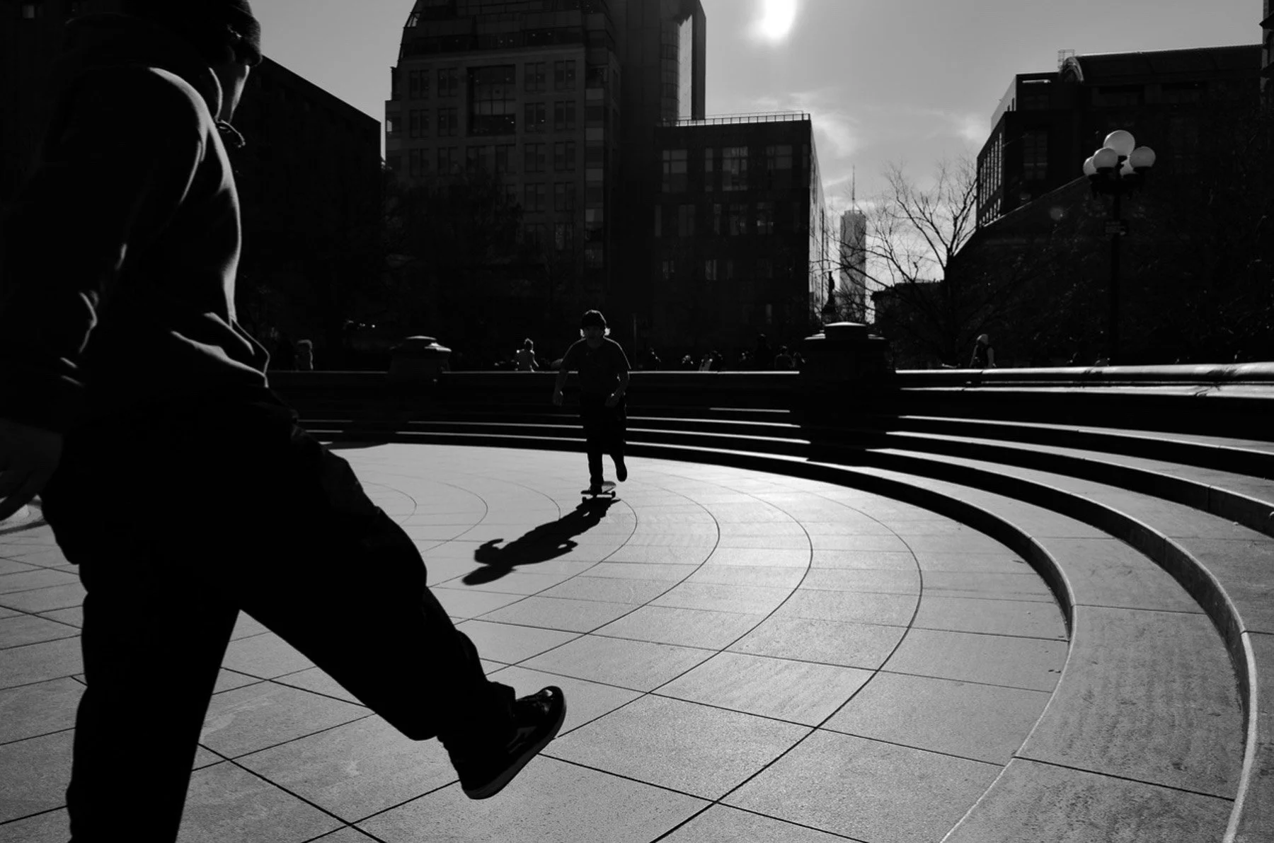 Black and white photo of a city park with a skateboarder in the middle, people sitting and walking in the background, tall buildings, and a partly cloudy sky.