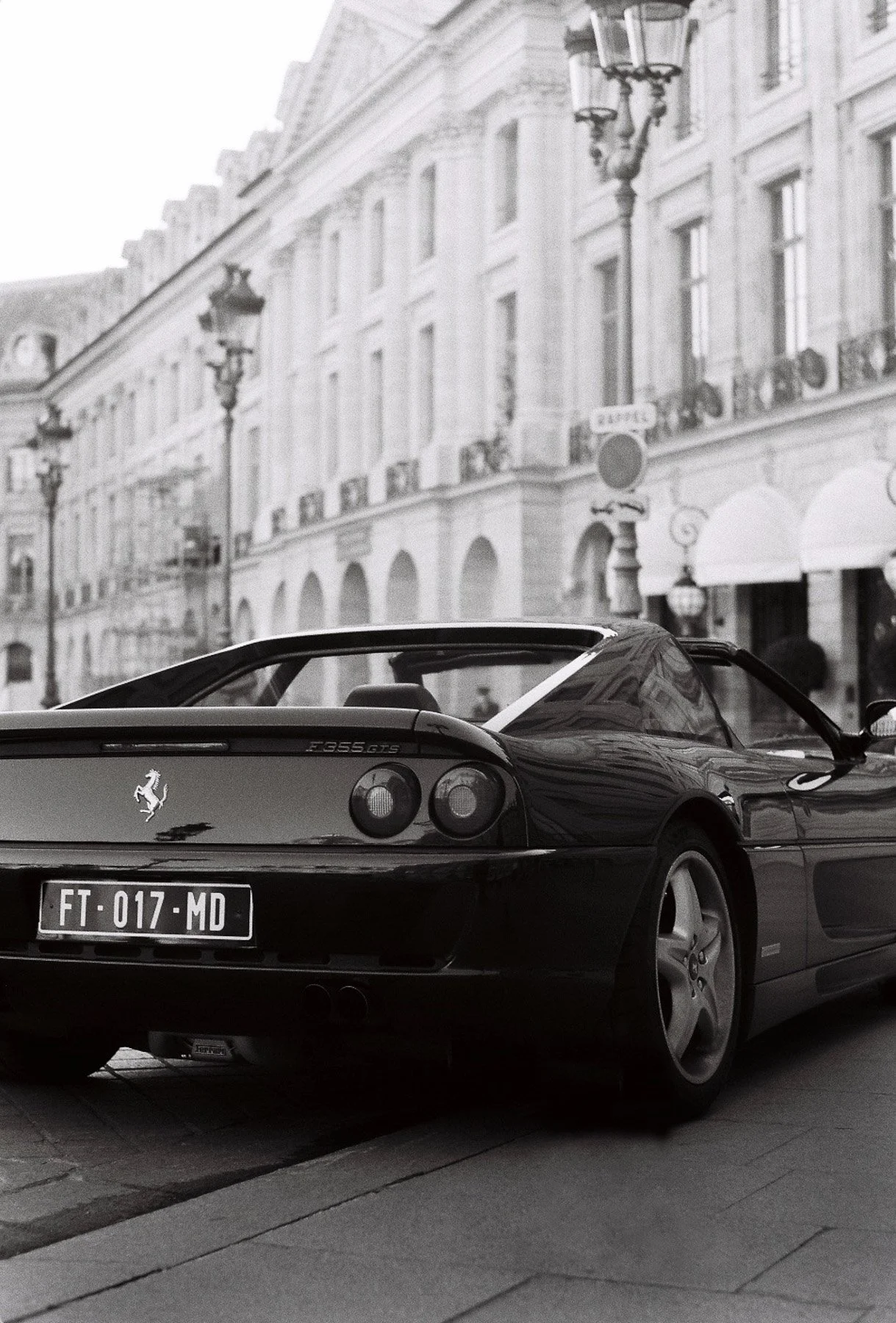 A black Ferrari F355 GTS parked on a city street with historic European-style buildings and street lamps in the background.
