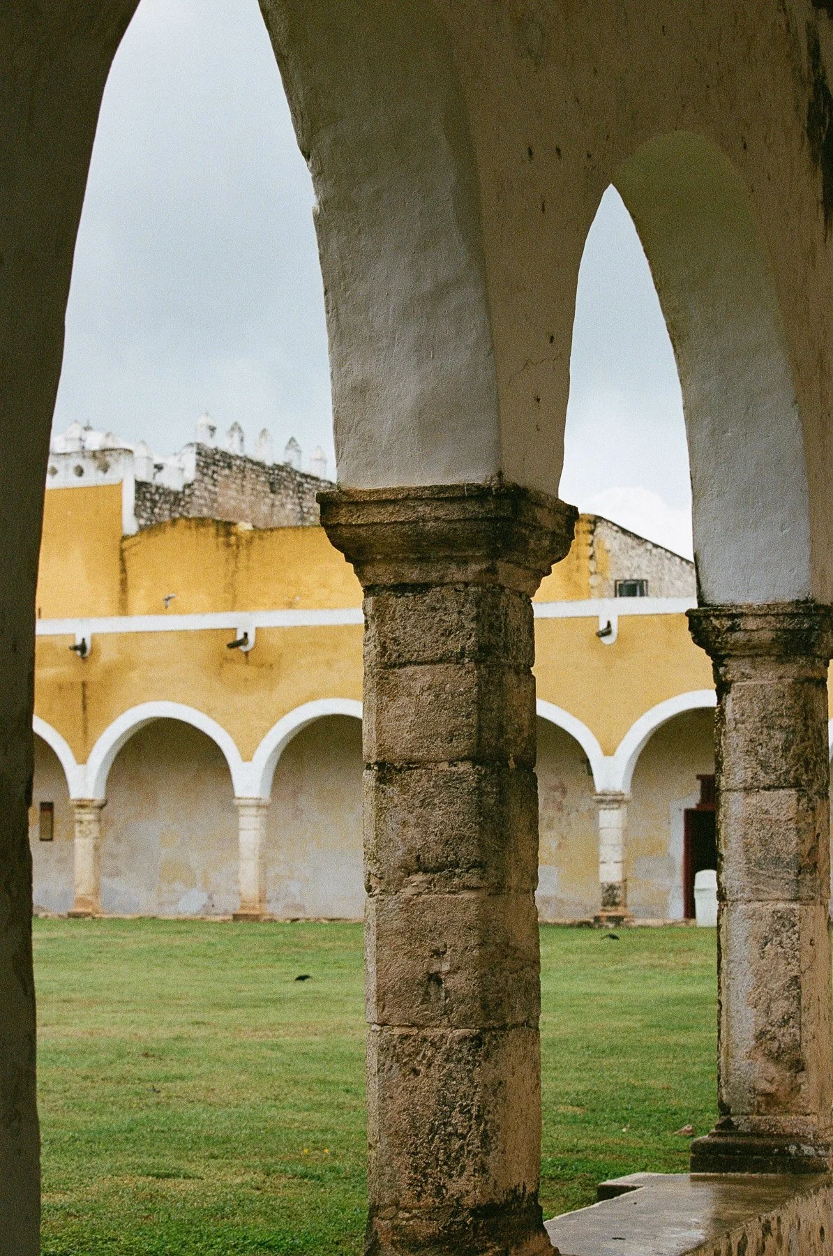 Ancient stone columns in a courtyard with a grassy area, archways, and a yellow building in the background.