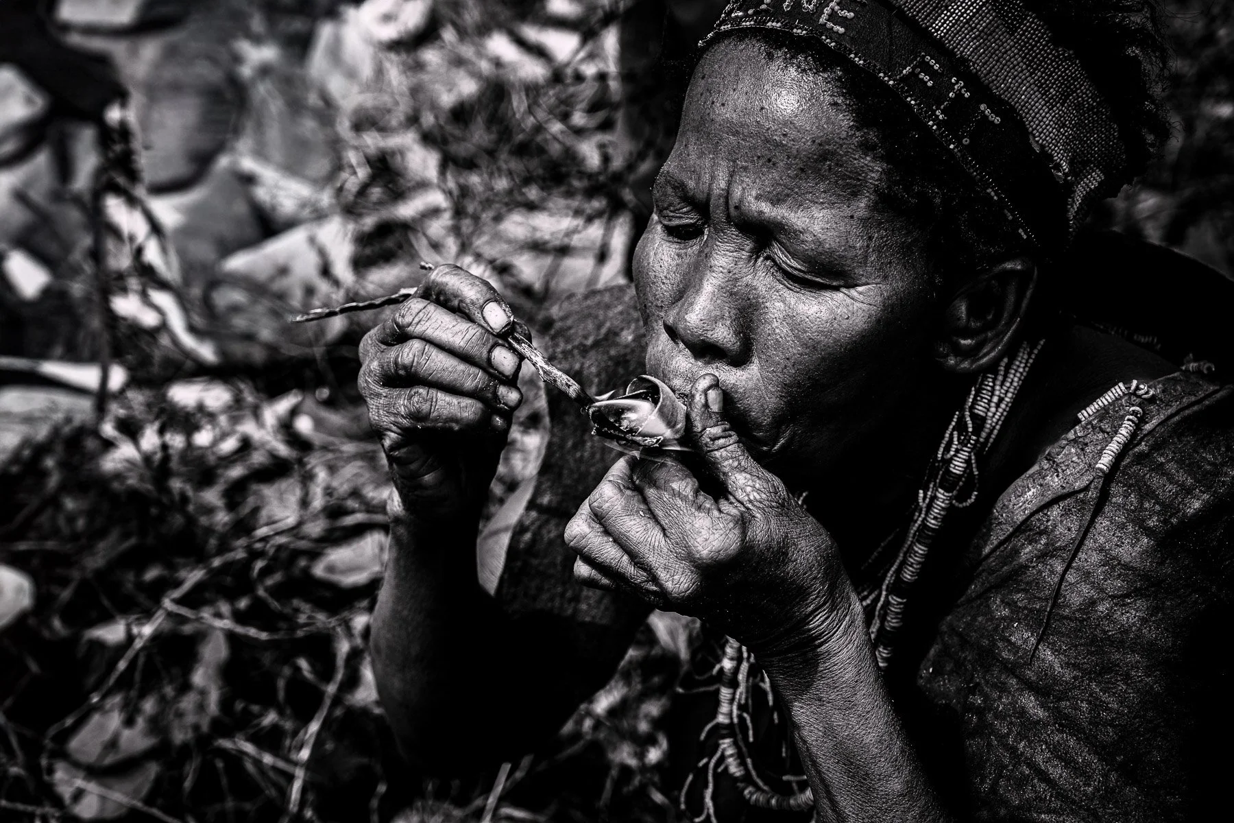 A woman with traditional jewelry and headwear, sitting outdoors and lighting a small object in her hands.