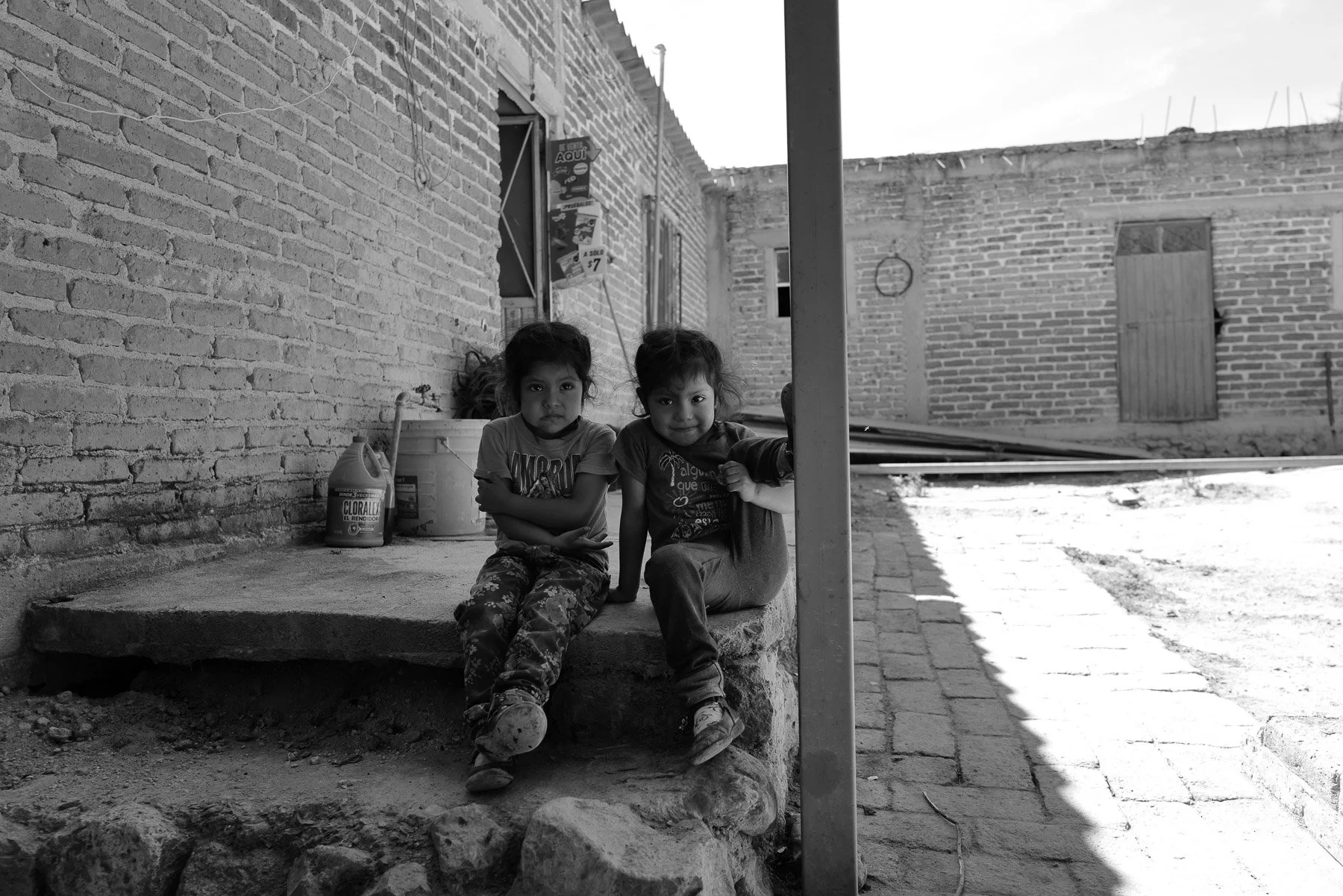 Two young children sitting on a concrete step outside a brick building, with supplies nearby, in a neighborhood with unfinished and simple structures.