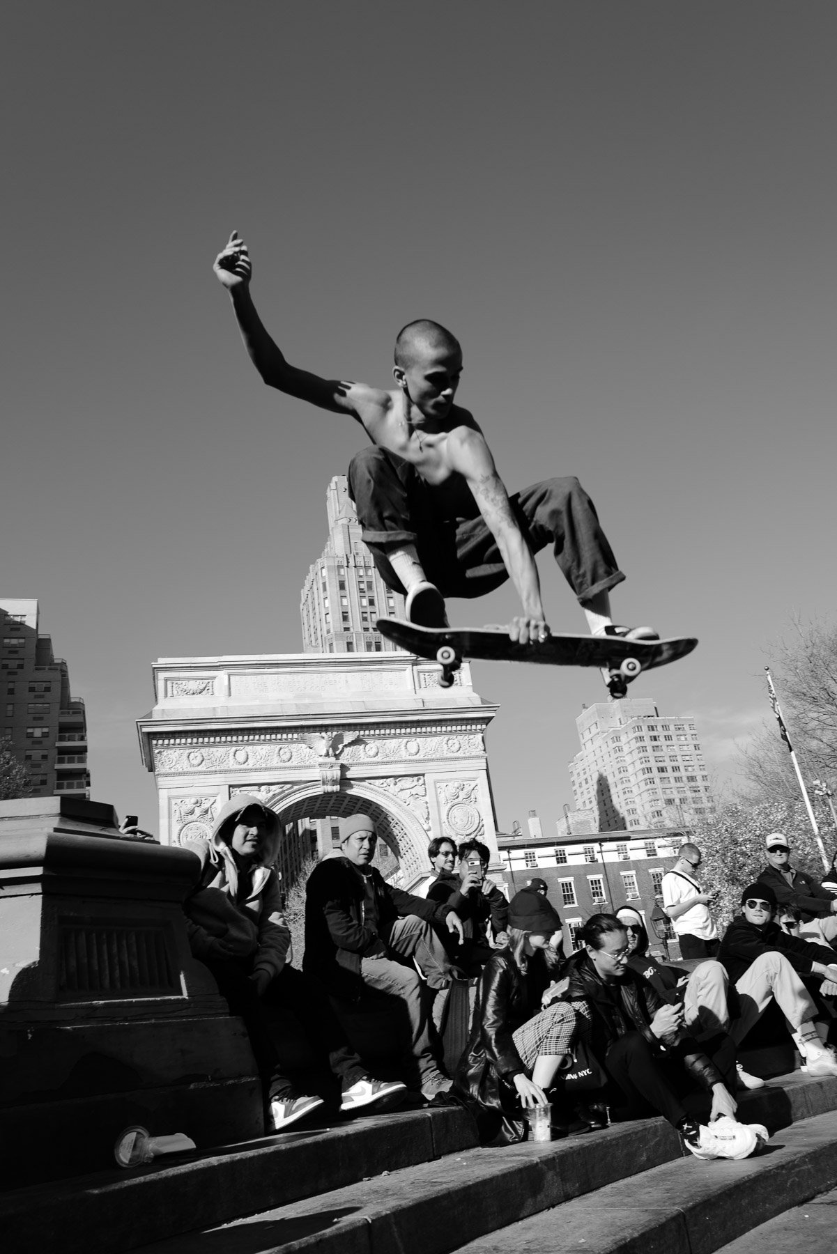 A young man skateboarding in a city park, mid-air with a skateboard beneath him, surrounded by onlookers sitting and standing on steps, with tall buildings and an arch structure in the background.
