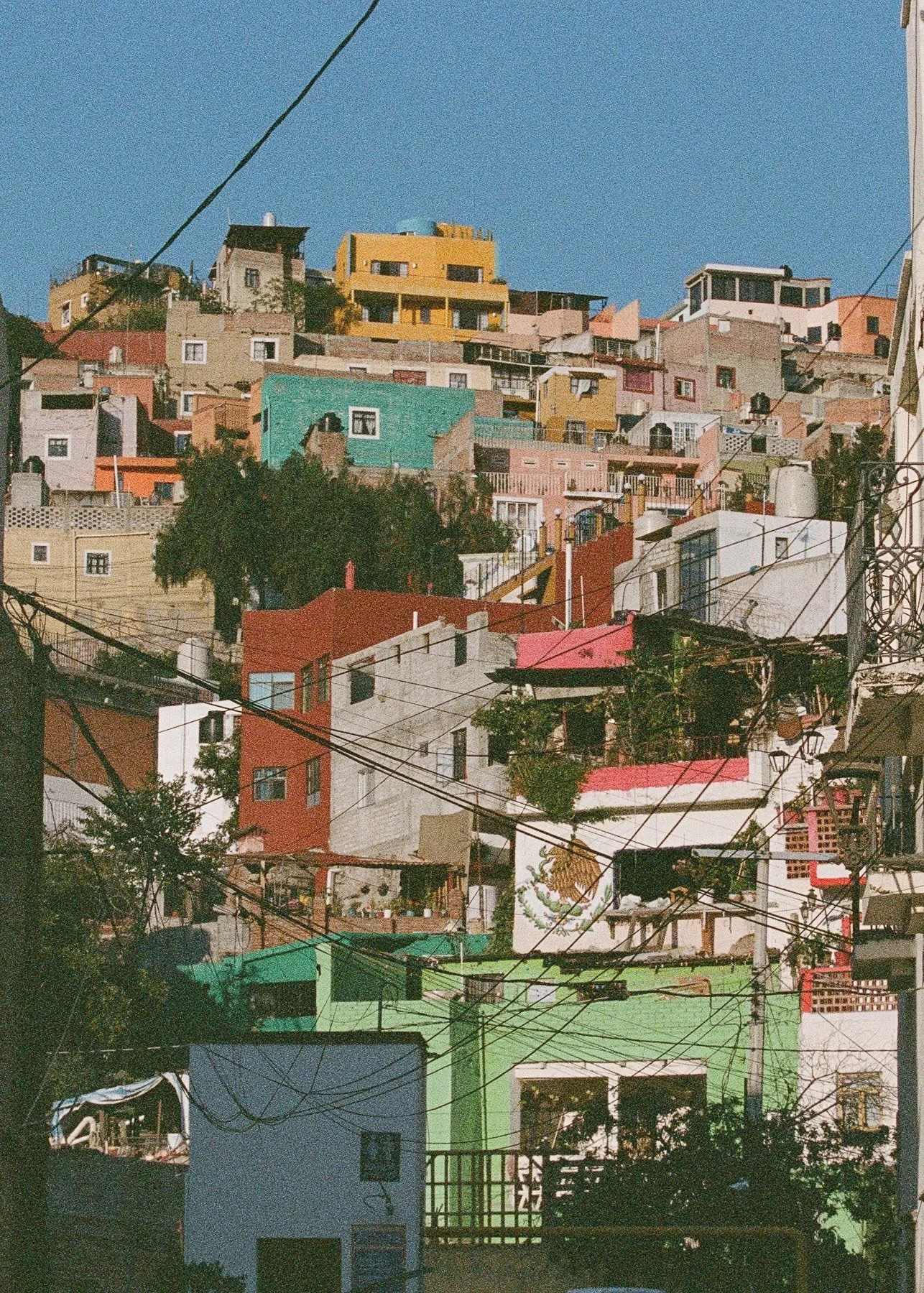 Colorful hillside neighborhood with variously shaped houses, some painted in bright colors like teal, yellow, and pink, stacked on a hill under a clear blue sky, with numerous power lines in the foreground.