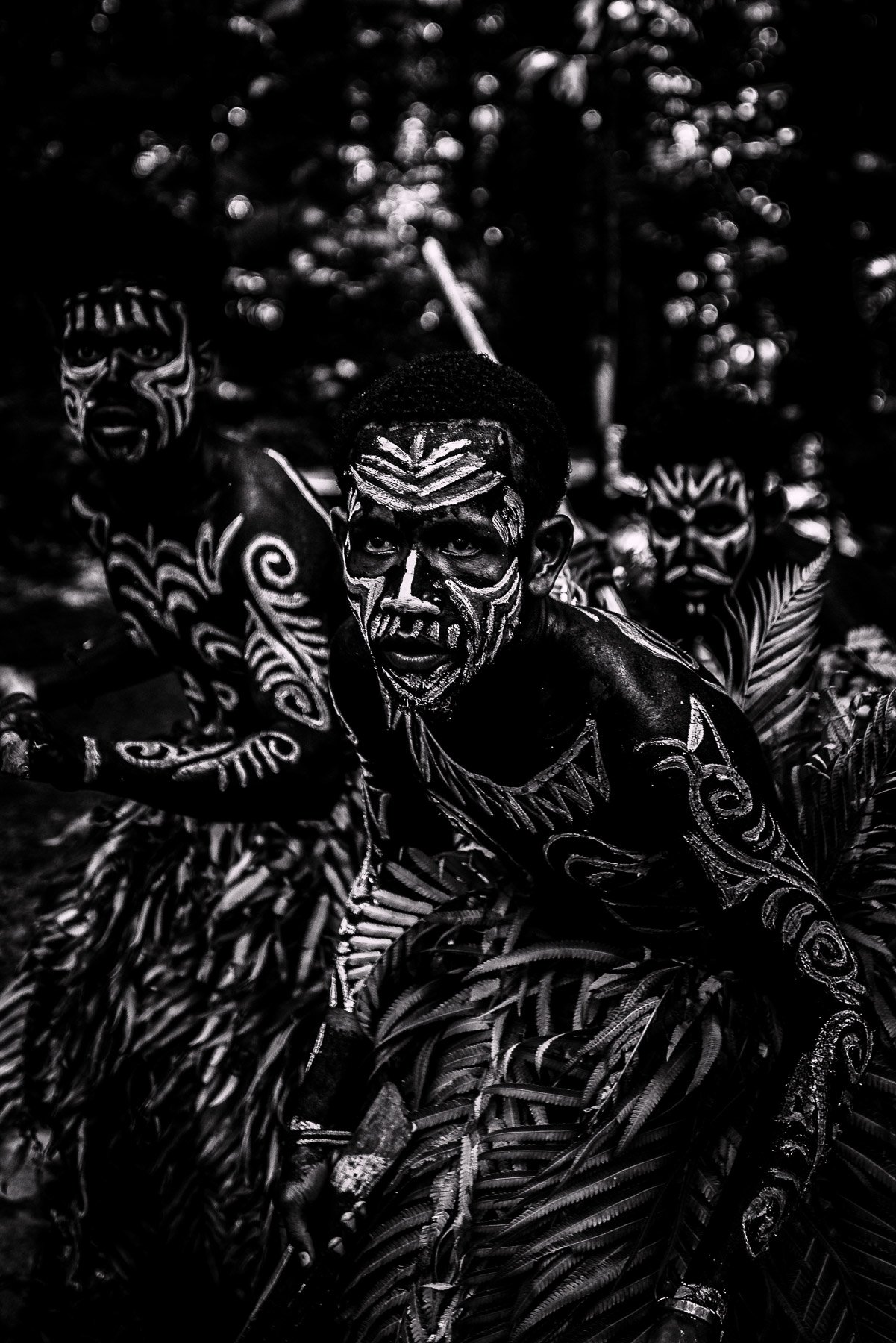 A group of men with traditional tribal face paint and body paint, dressed in patterned clothing, performing a cultural dance or ritual outdoors.