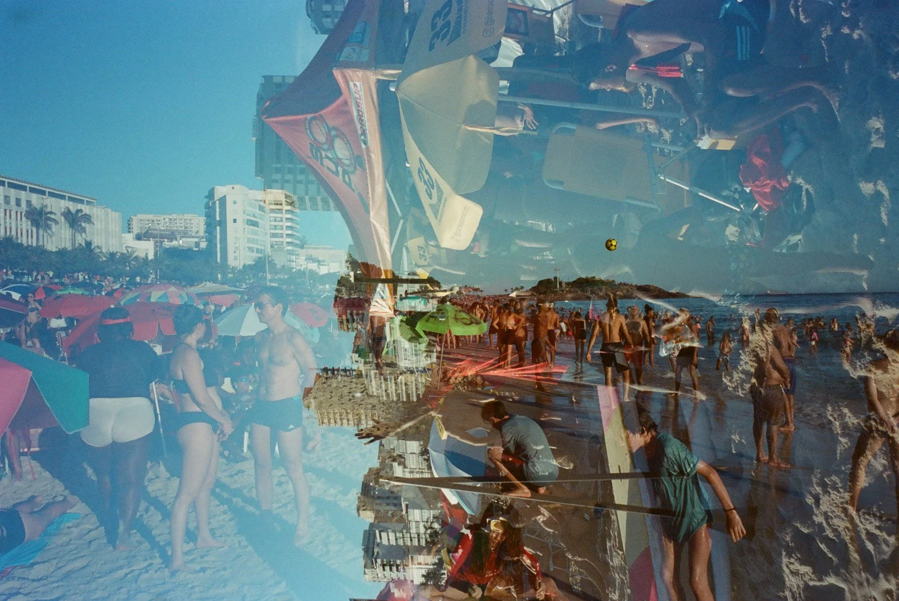 A crowded beach scene with many people in swimsuits under umbrellas near the water, with city buildings in the background, and some people playing with a ball in the ocean.
