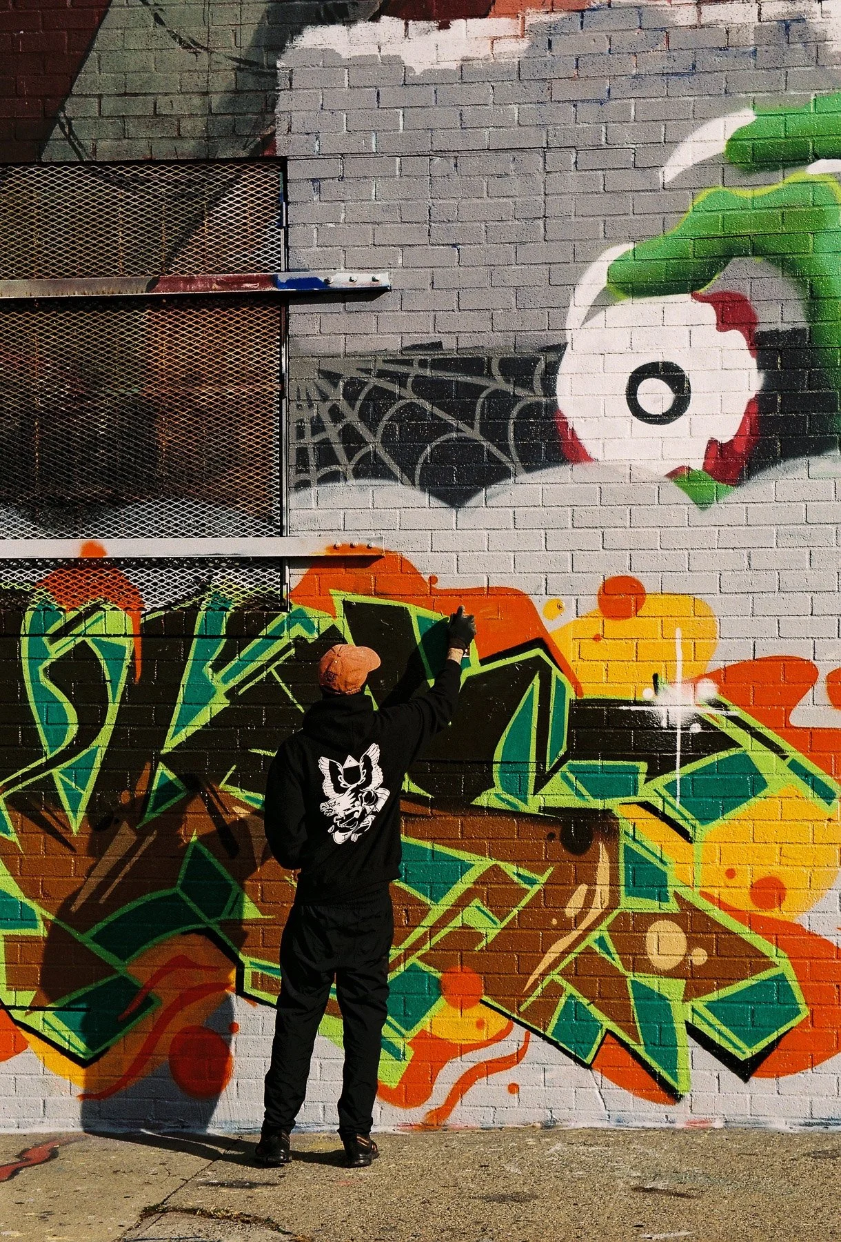 A person wearing a black hoodie with a fox design and orange hat is spray painting graffiti on a brick wall featuring colorful abstract shapes and a large stylized apple with a spider web design.