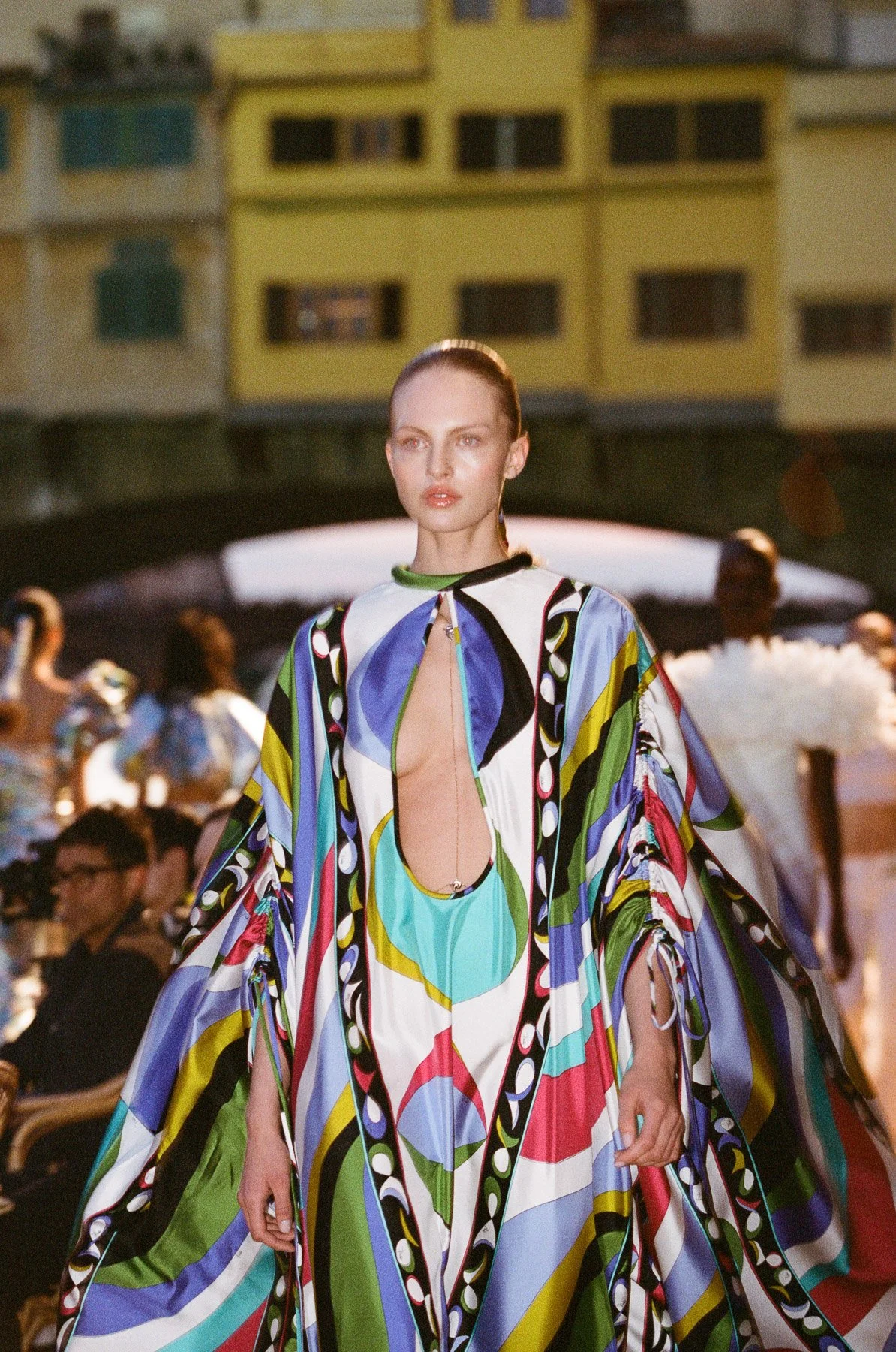A fashion model walking down an outdoor runway during a fashion show, wearing a colorful, patterned dress with a deep neckline. The background shows an audience and a cityscape with apartment buildings.