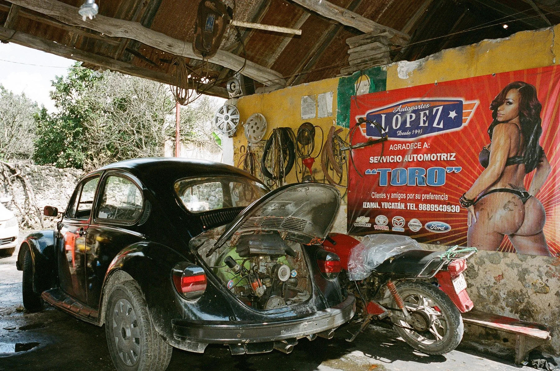 Inside a mechanic's garage with a vintage black Volkswagen Beetle and a motorcycle. A large red and orange banner advertises 'Autopartes López' auto parts, featuring a woman in revealing clothing. The garage has tools hanging on the yellow wall and a
