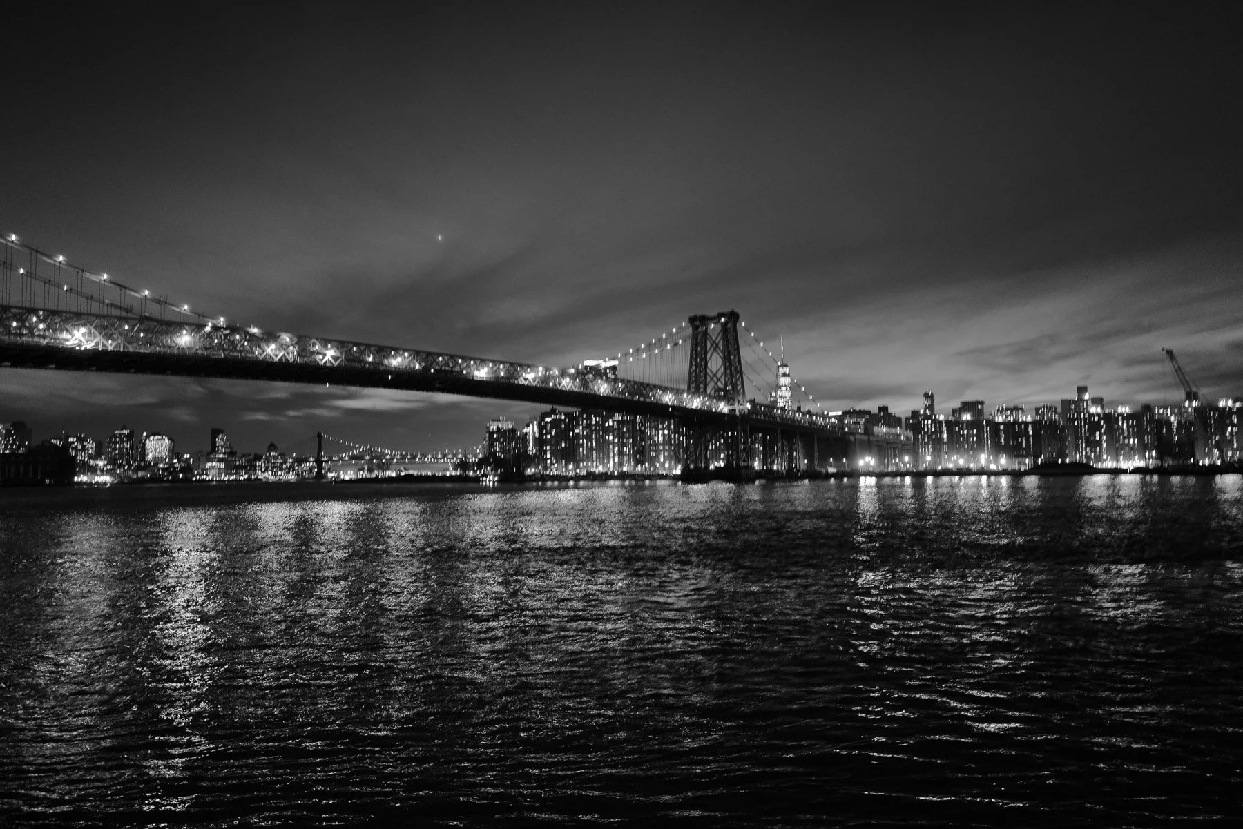 Black and white photo of a large suspension bridge over a body of water at night with a city skyline in the background.