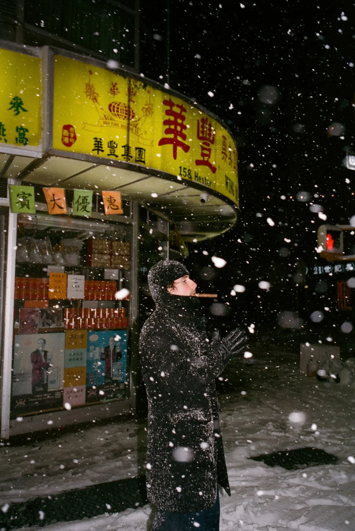 Person in black winter coat and gloves standing outside in heavy snowfall, smoking a cigar, near a store with Chinese signage and posters in an urban area at night.