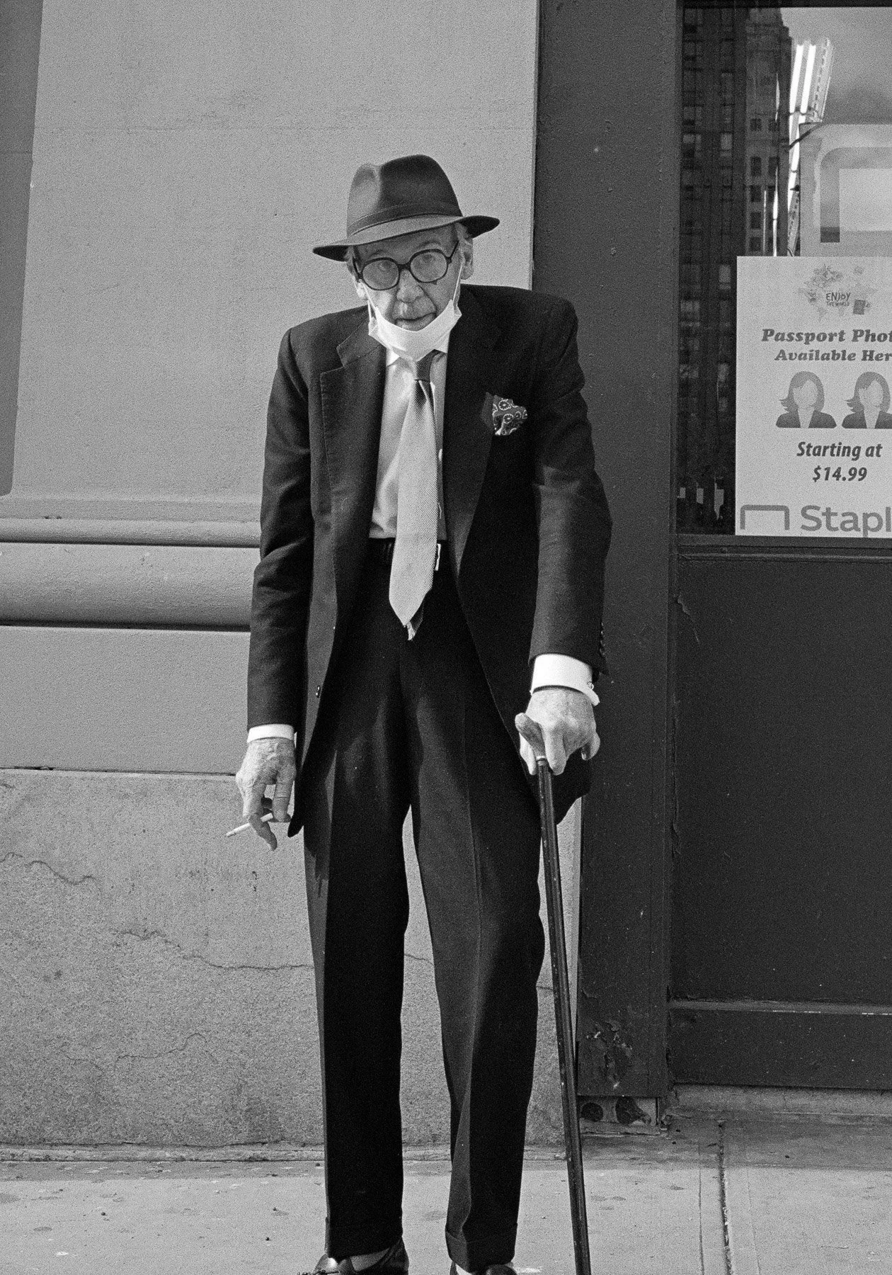 A black and white photo of an elderly man wearing glasses, a fedora, a face mask, a suit, tie, and a pocket square, standing on a city sidewalk with a cane and holding a cigarette.