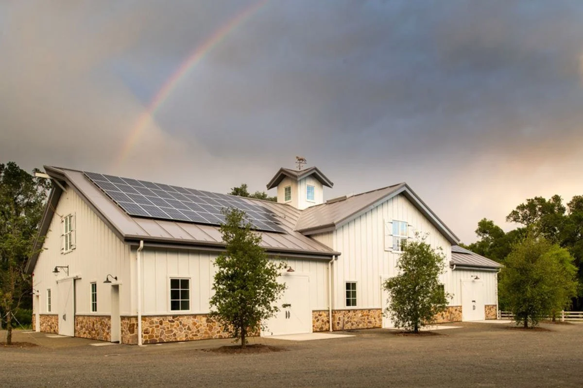 White barn with solar panels on the roof, surrounded by trees, and a rainbow in the sky.