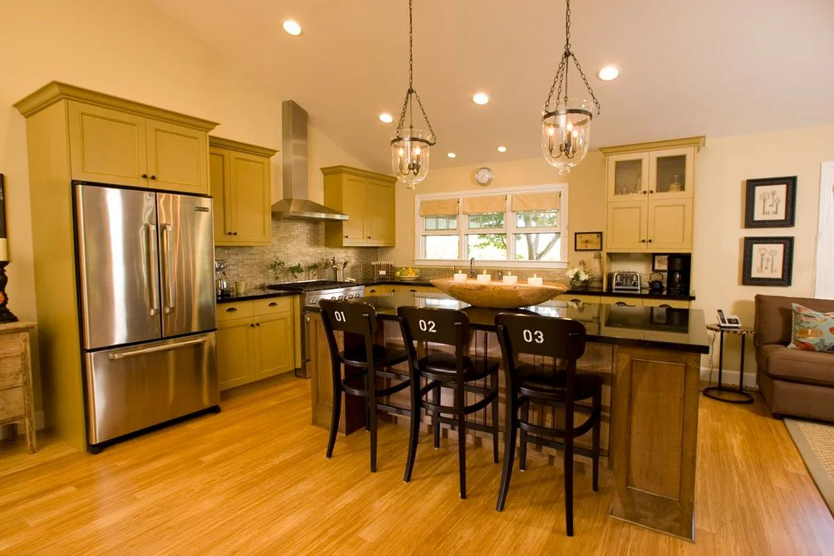 Kitchen with yellow cabinets, stainless steel refrigerator, black countertops, three black chairs with numbers 01, 02, 03, a window with three panes, and a kitchen island with candles.