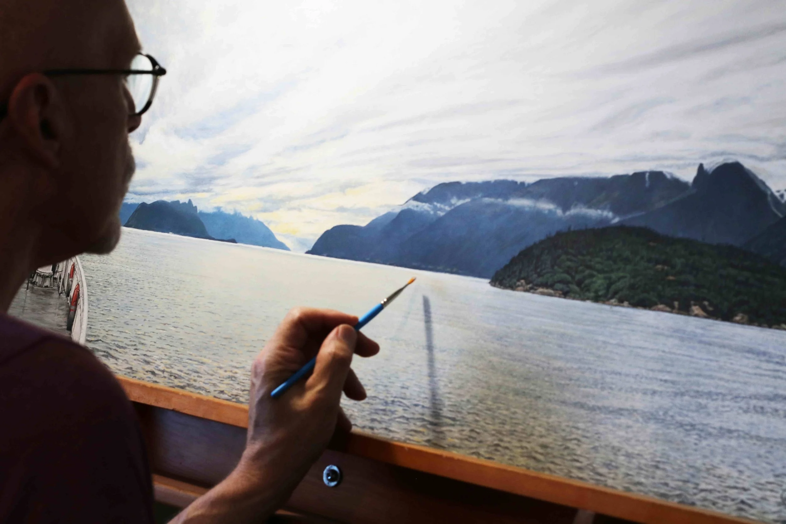 An artist painting a landscape of a lake with mountains in the background from a boat or dock, holding a paintbrush near the painting.