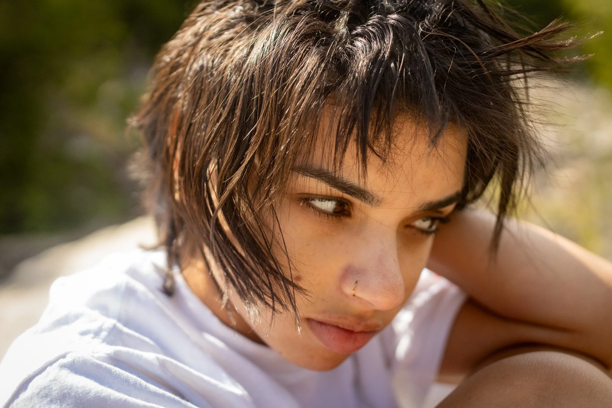 Close-up of a woman with wet, tousled dark hair, light skin, brown eyes, a small nose ring, and wearing a white shirt, looking downward outdoors.