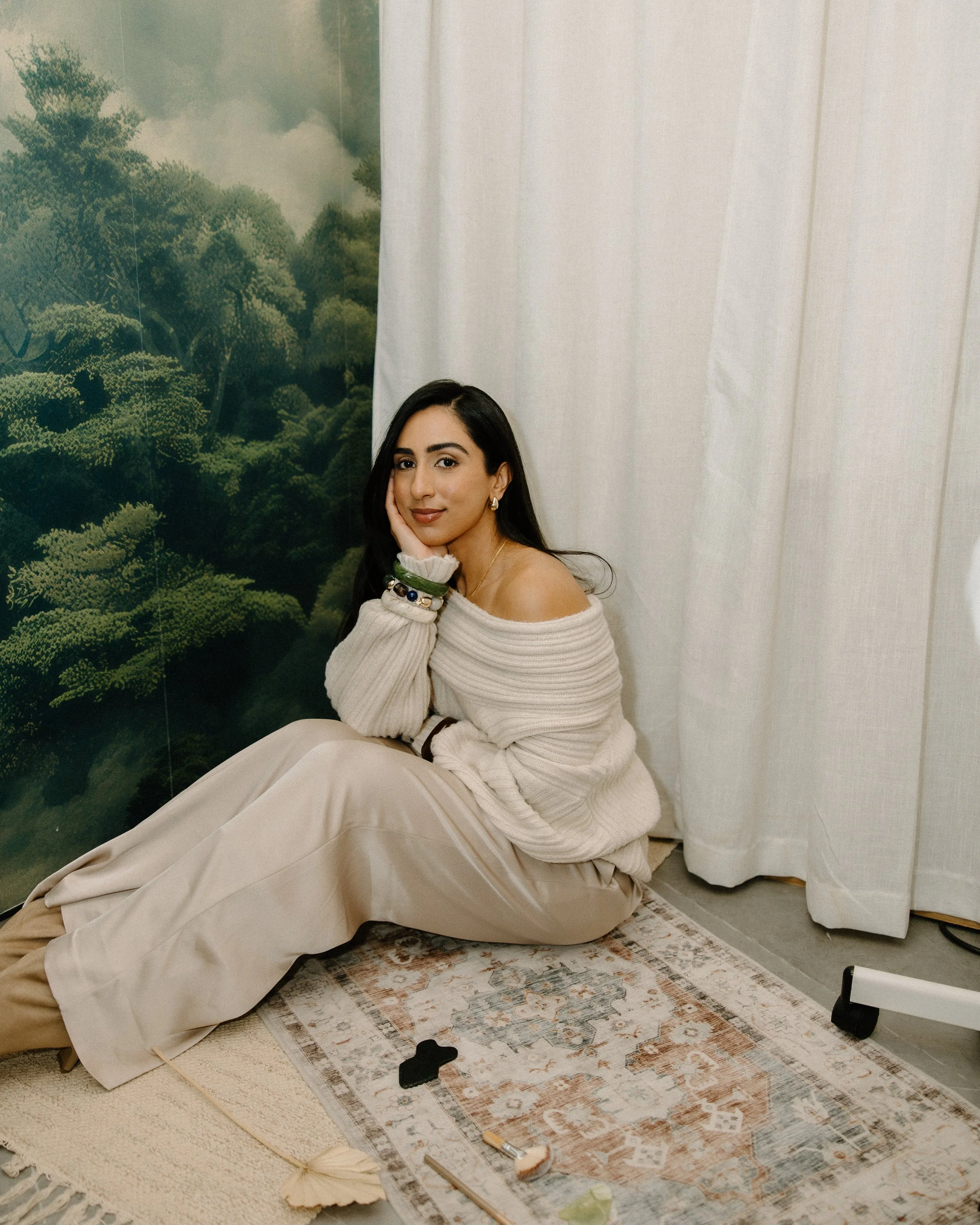 A woman with dark hair and tan skin sitting on the floor, wearing a beige off-the-shoulder sweater and beige pants, resting her head on her hand, with a serene expression. Behind her is a backdrop of green trees and white curtains, with a patterned rug under her.