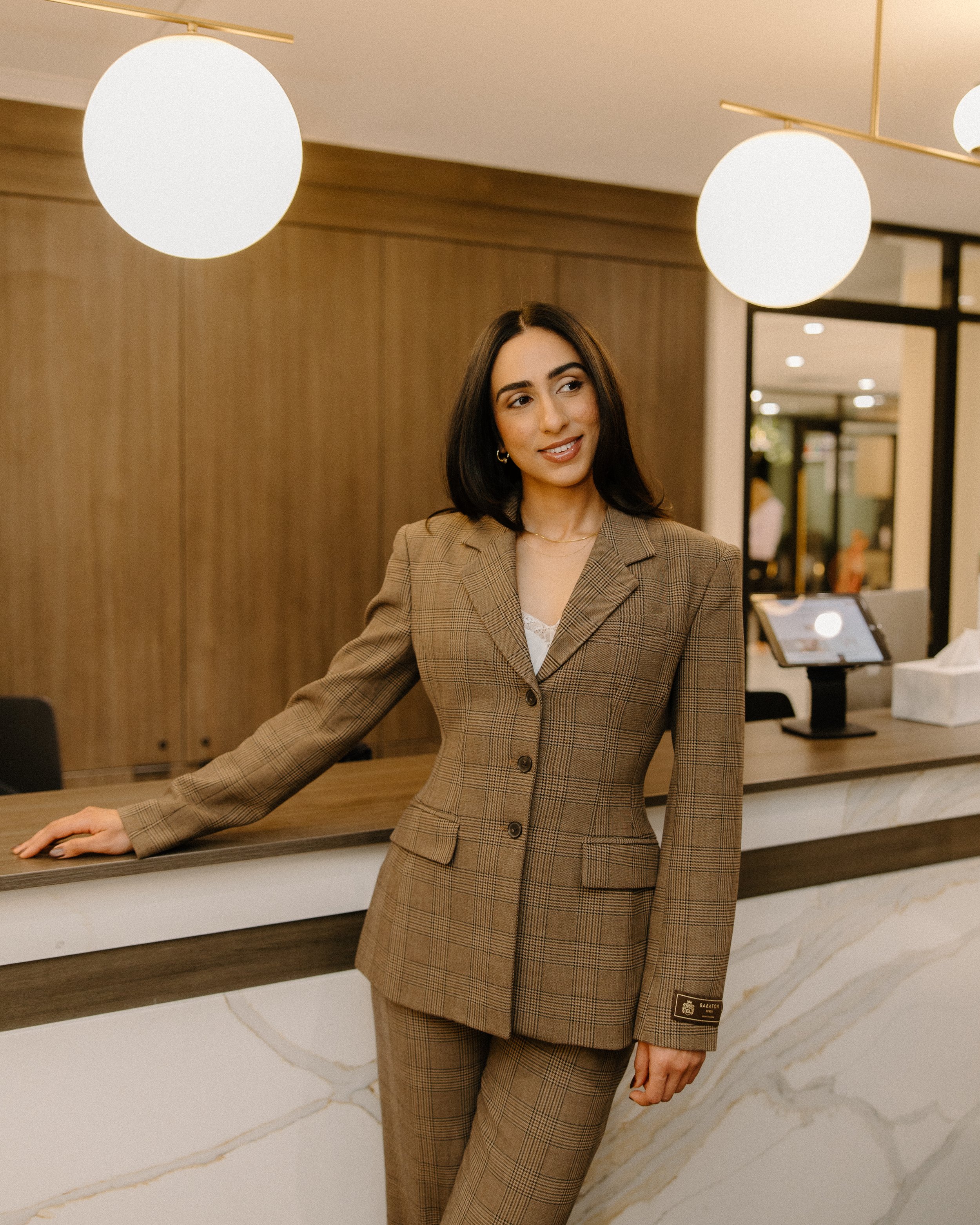 A woman with dark hair in a checkered brown suit standing at a reception desk in a modern hotel lobby.